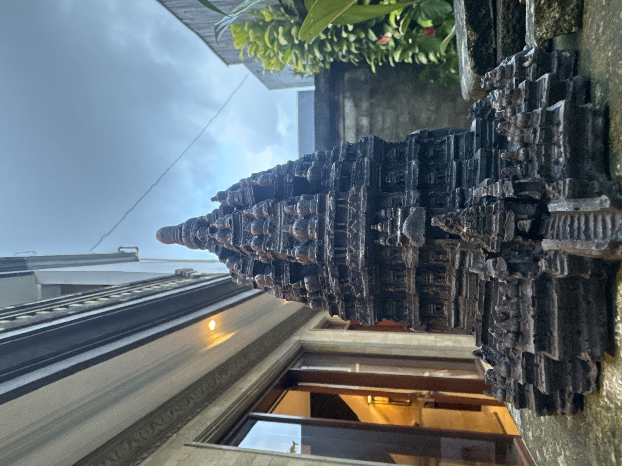 An outdoor, upward-angled shot in Ubud District, Indonesia, captures a highly detailed miniature temple or stupa-like structure, likely made of dark, weathered stone or carved wood, in the foreground. The intricate, multi-tiered edifice rises to a pointed spire, adorned with numerous architectural decorations, small alcoves, and what appear to be tiny sculpted figures or relief carvings integrated into its layered design. The overall aesthetic is reminiscent of traditional Southeast Asian temple architecture.
In the mid-ground and background, a modern building with a light-colored facade is visible, featuring windows through which an interior artificial light source glows warmly. Above and to the left of the building, green foliage from plants or trees adds a natural element to the scene. The sky above is overcast with soft, diffused clouds, through which the sun faintly illuminates the scene. The perspective suggests the miniature structure is placed on a ledge or wall, with the building and natural elements forming its backdrop. There is no discernible activity taking place, and no people are visible. The image has a slightly tilted orientation, suggesting it might have been taken with the camera rotated. No visible text is present.
