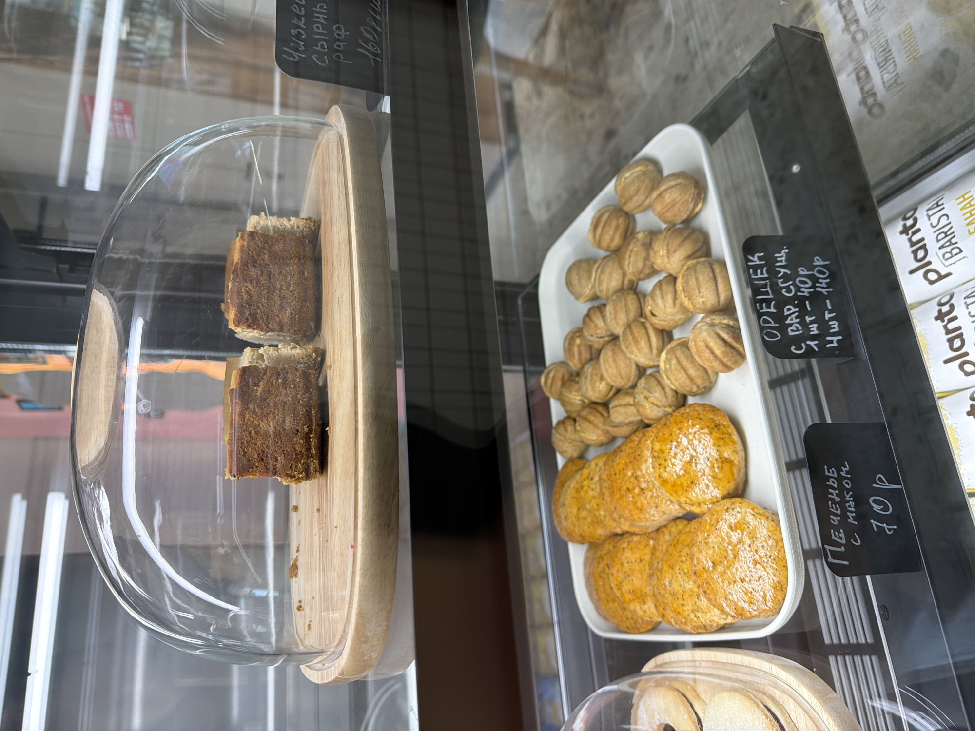 The image shows a display case in what appears to be a food establishment, likely located in the city of Chaikovsky, Russia, based on the Cyrillic text on the price tags. Inside the case are several types of pastries. On the left, under a clear dome, are two slices of a dark-colored cake or bread, displayed on a wooden board. To the right of that is a white rectangular tray containing two types of pastries: small walnut-shaped cookies and round poppy seed cookies. The poppy seed cookies are labeled at 70 Russian Rubles. A sign for the walnut-shaped cookies indicates that they are filled with condensed milk and cost 40 Rubles each, or 140 Rubles for a pack of four. The cake under the dome is also labeled, but the beginning of the word is illegible, although it is clear that it is a cheesecakes or something similar, and sells for 160 Russian Rubles. To the right, partially visible, are packages of "Planto Barista" plant-based milk for making coffee or tea. The display case itself is made of glass and black metal. The background includes a blurred view of the interior, possibly a window reflecting the outside environment.