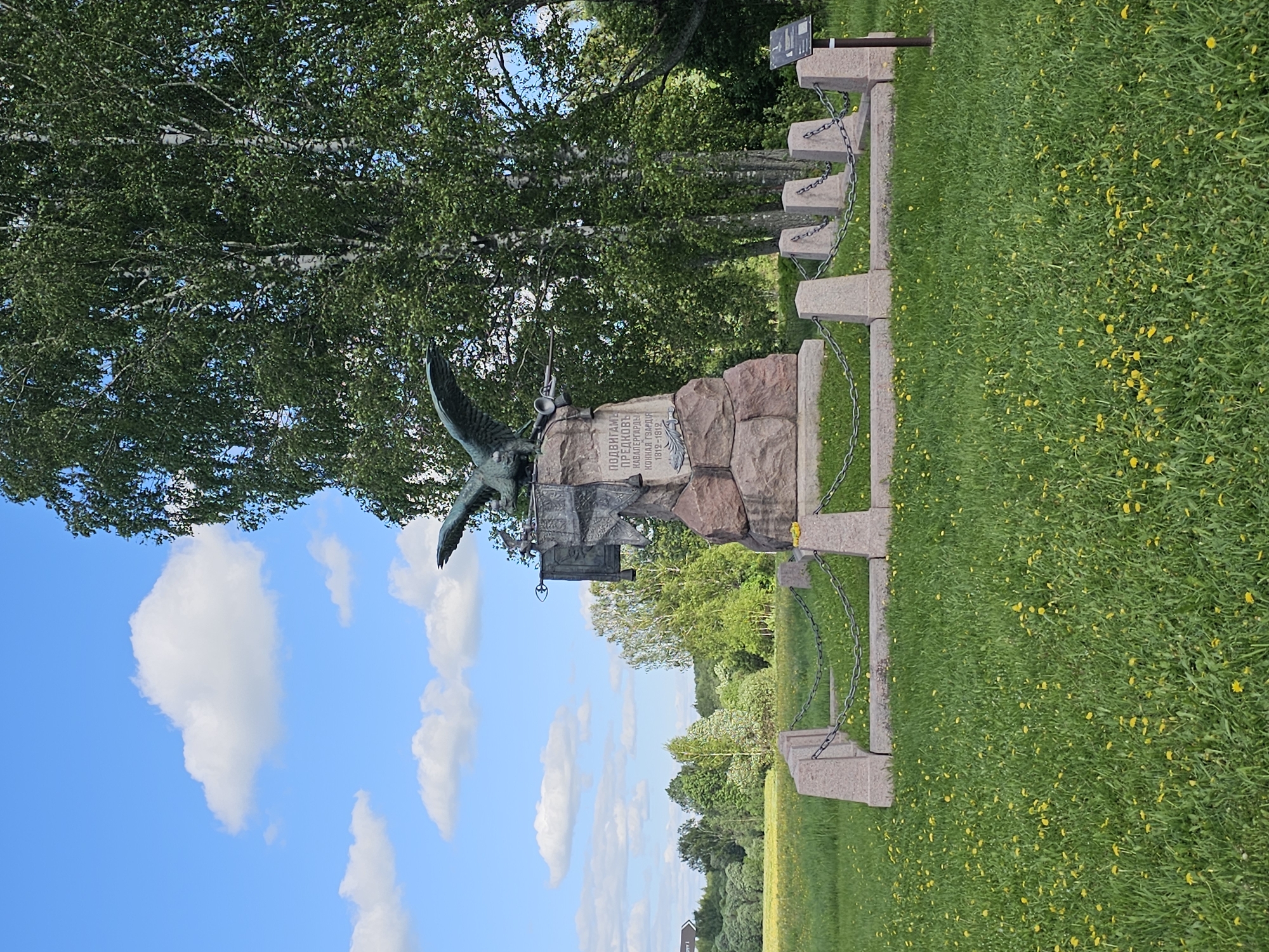 The image depicts a monument dedicated to the Cavalry Guard Regiment located in the city of Borodino, Russia. The monument is composed of a bronze eagle perched atop a stone pedestal, with an inscription on the pedestal in Russian. A chain-link fence surrounds the monument, and lush green grass with yellow flowers covers the ground. A dense canopy of trees frames the monument, while a blue sky dotted with fluffy white clouds provides the backdrop.