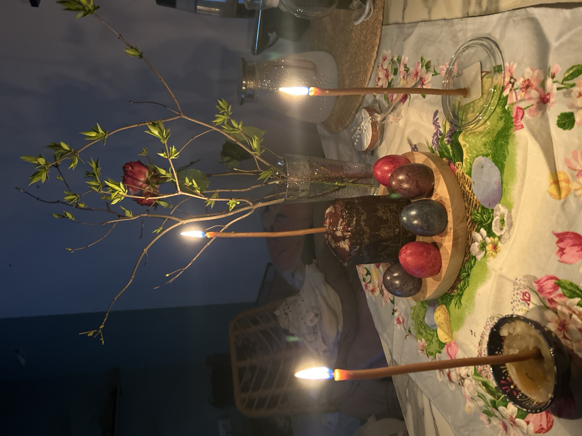 The image depicts a table set with what appears to be items for an Easter celebration. There is a decorated Easter cake, several dyed eggs, and lit candles. There is a vase of budding branches with a single rose. The table is covered with a floral patterned tablecloth. The location is unknown, however it could be in Russia based on the Easter-related objects.