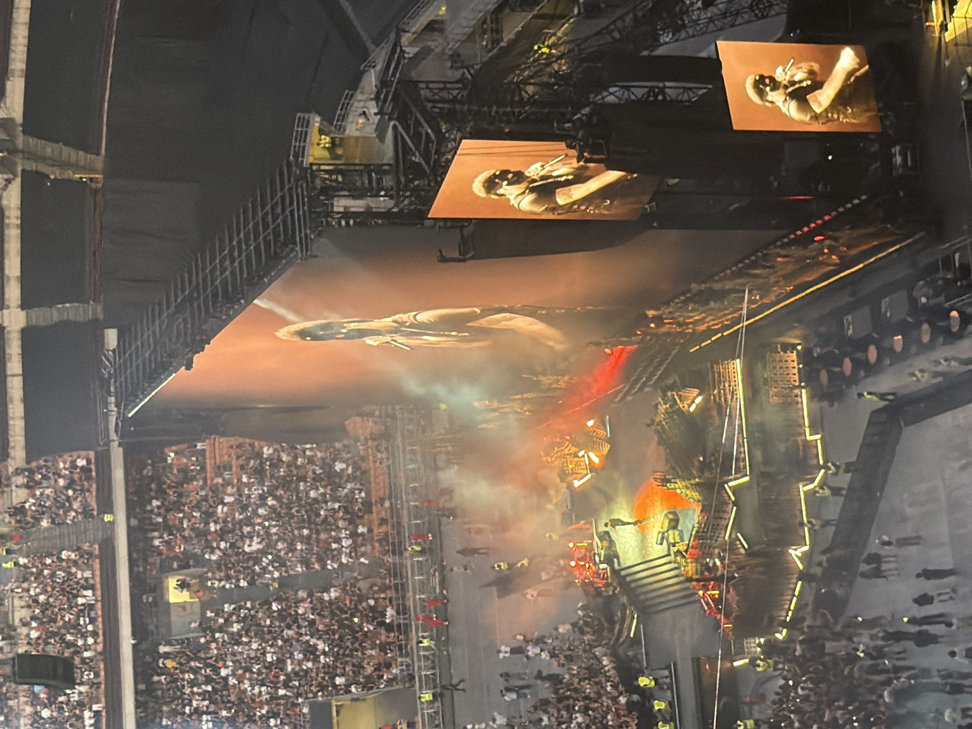 An aerial, high-angle, wide shot captures a live concert at San Siro Stadium in Milan, Italy. The stadium is packed with an audience, with rows of spectators visible in the background. A large screen displays a performer singing into a microphone, and there are additional screens showing close-ups of the performer. The stage is lit with dramatic lighting, featuring orange and red hues, and smoke effects are present. There are crew members and performers on stage, interacting with the setup. The overall scene depicts a high-energy live musical performance.