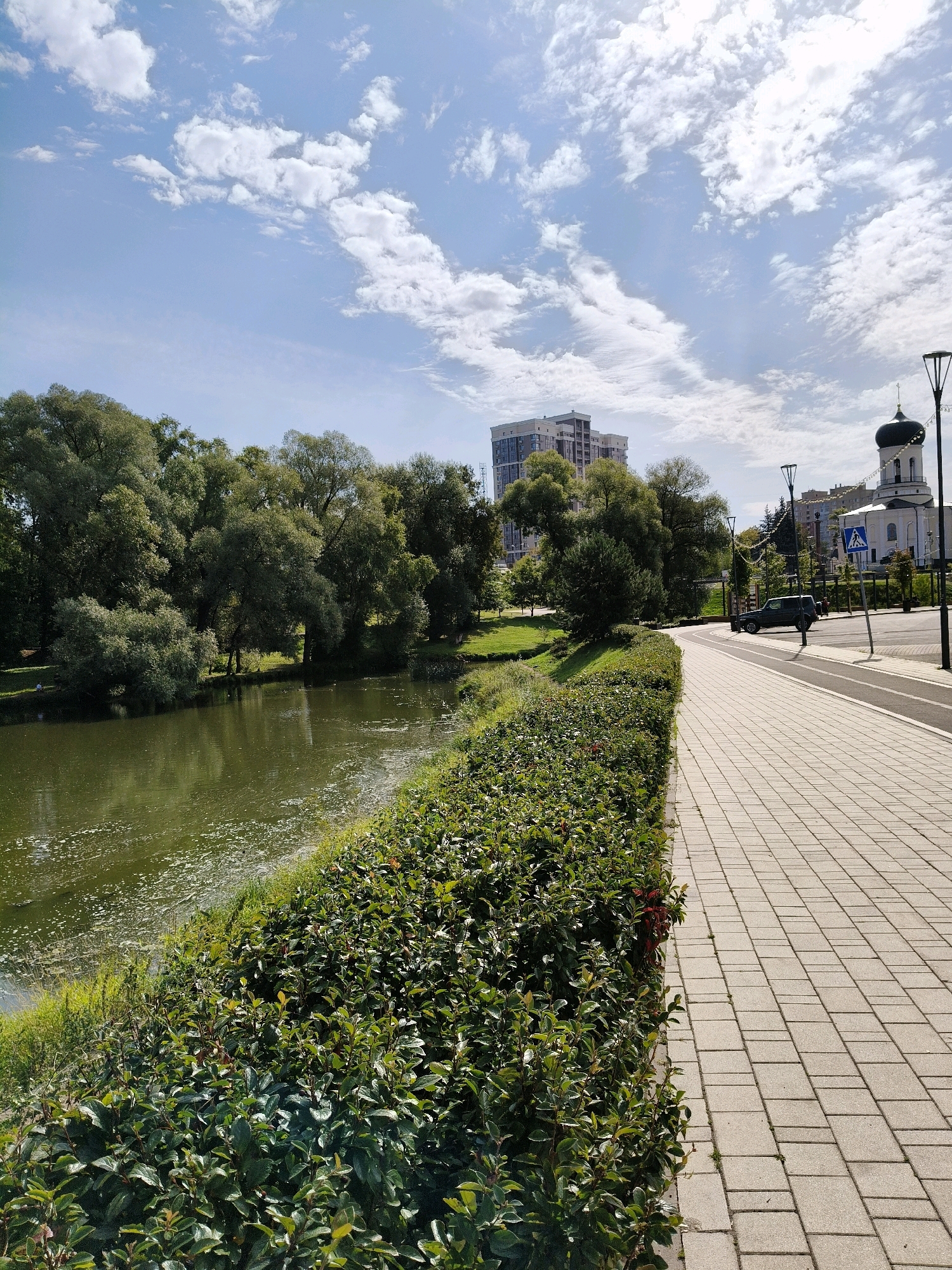 The image shows a waterfront promenade in Naro-Fominsk, Russia, on a sunny day with scattered clouds. A paved walkway with square tiles runs along the right side of the frame, bordered by a manicured green hedge. Across the walkway, a dark-colored SUV is parked on the side of a road. Further down the road, a white church with a dark dome and a cross stands prominently, with other buildings visible in the background, including a tall, modern residential building. On the left side of the walkway, a body of water, likely a river or pond, is visible, with lush green trees lining its bank. The water's surface has some foam or debris. A few small figures, appearing to be people, are present in the distance near the trees by the water. The overall scene depicts a public urban space that blends natural elements with modern infrastructure and historical architecture.