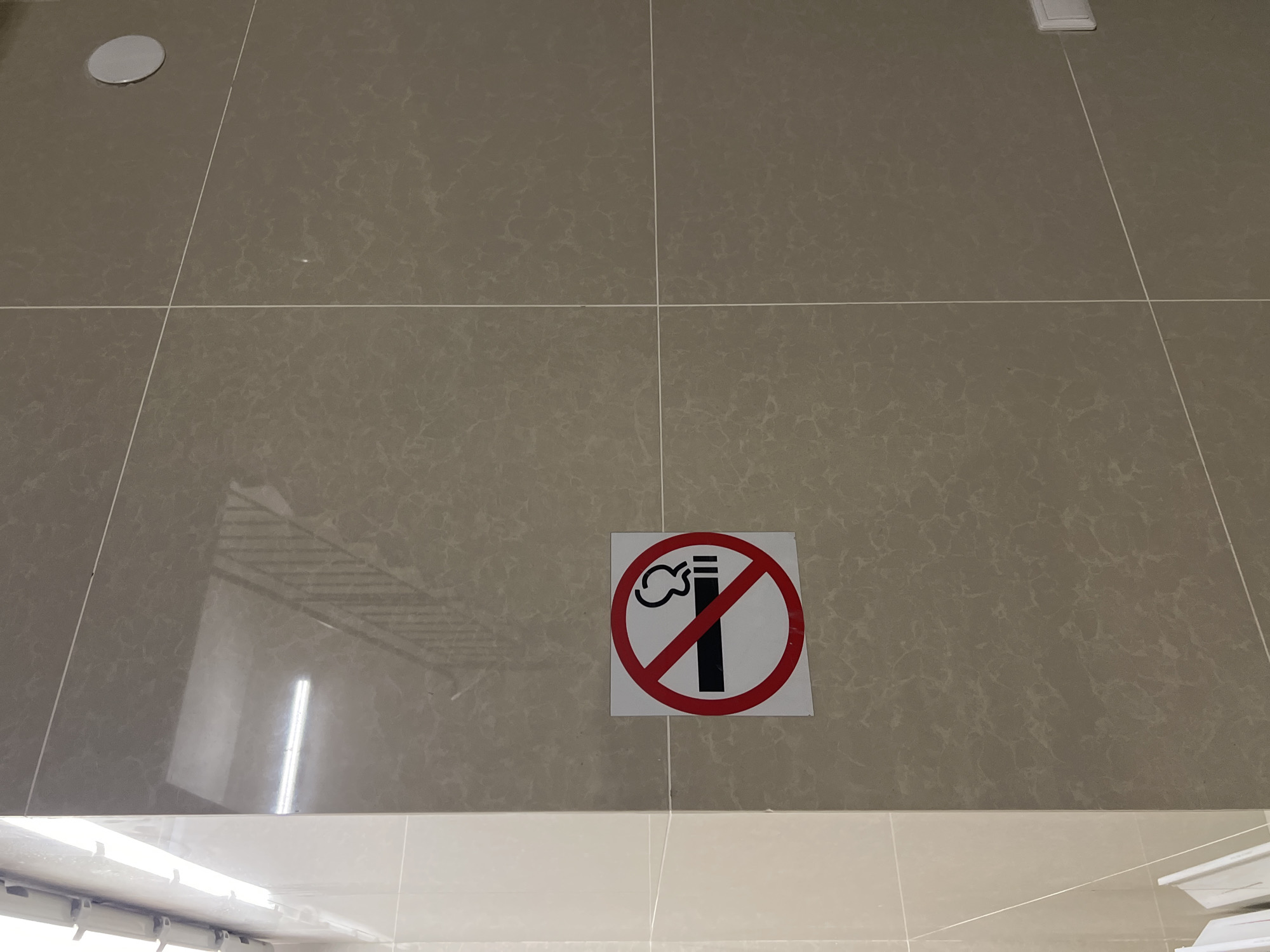 The image shows a section of an interior wall covered in large, square, light brown tiles with a subtle patterned texture. A "No Smoking" sign, circular with a red border and a diagonal red line through a cigarette symbol, is affixed to the tiles. The sign is white with black and red graphics. To the left, the wall transitions to a lighter, cream-colored tiled surface, with a fluorescent light fixture visible at the top. Reflections of a staircase with railings are visible on the tiled surfaces. A white circular object, possibly a speaker or light cover, is mounted on the upper right section of the wall. A white electrical outlet is visible on the lower right side of the wall. The scene appears to be an indoor public space, such as a hallway or stairwell. The location is not identifiable as Krasnoyarsk, Russia, or any other specific city.