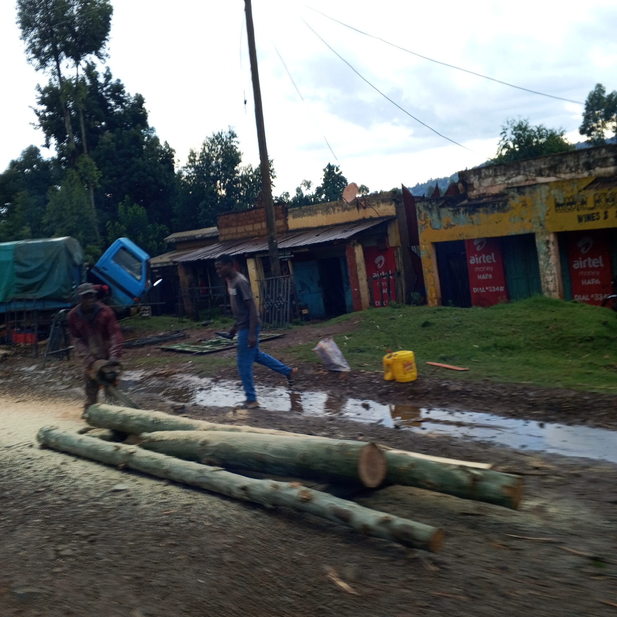 The scene depicts a street in Borabu Chitago ward, Kenya, after rainfall, with puddles on the unpaved ground. In the foreground, three logs lie amidst sawdust, indicating recent cutting activity; a man operating a chainsaw is visible on the left. Another man is walking past the logs, away from the chainsaw operator.
Behind them, a truck with a blue cab sits askew. The backdrop features a row of dilapidated buildings with advertisements for "Airtel Money Hapa" and "Wines &". The buildings are painted in yellow and red, but the paint is peeling. Beyond the buildings, trees are visible. The sky is overcast, suggesting recent or ongoing rain.