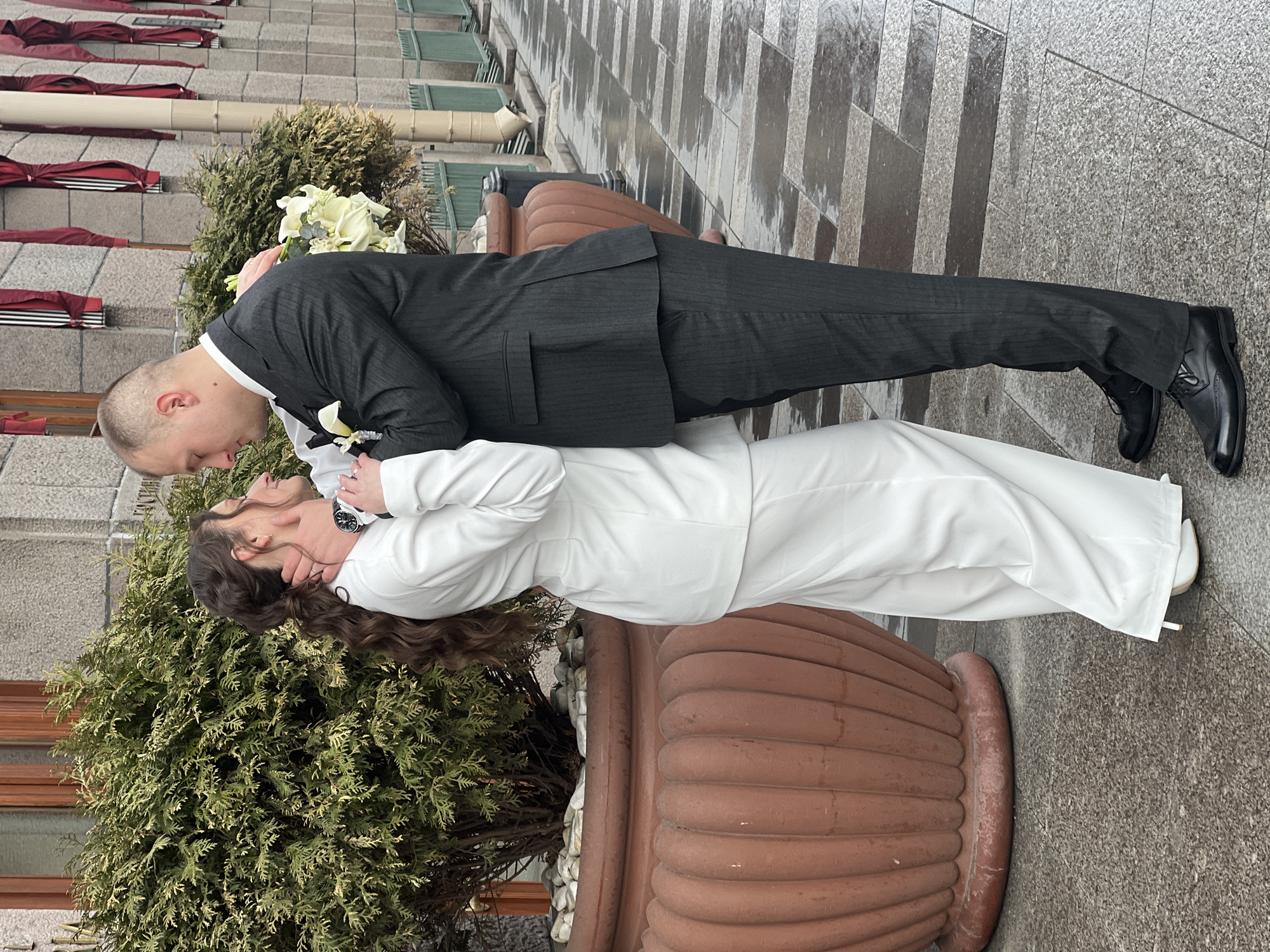 A man and a woman are embracing each other. The man is wearing a dark suit and the woman is wearing a white suit. They are positioned next to a large planter with shrubs in it. The scene is located on a gray stone walkway near a building. The building has a gray facade, windows, and awnings. The location is Saint Petersburg, Russia.