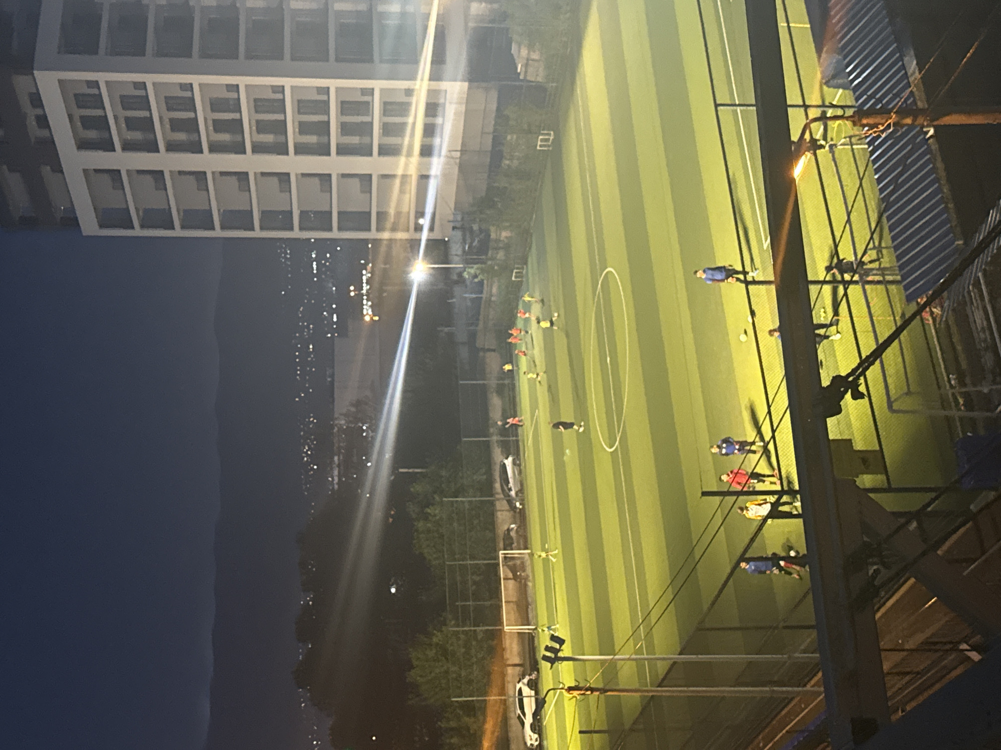 The image depicts an outdoor soccer field illuminated by artificial lights at night in Tbilisi, Georgia. A modern, multi-story building with a light-colored facade and dark-framed windows dominates the right side of the frame, casting a shadow over part of the field. The soccer field itself is artificial turf with visible yard markings. Several individuals, appearing to be a mix of adults and children, are spread across the field, engaged in what appears to be a soccer game or practice. Some are wearing athletic clothing, and a soccer ball is visible on the field. In the background, a cityscape with scattered lights suggests a populated area, and distant mountains are silhouetted against the dark sky. The foreground shows a section of a structure with a corrugated roof and visible metal beams, possibly a viewing area or part of an adjacent facility. A street lamp is also visible in the foreground, adding to the nighttime illumination.