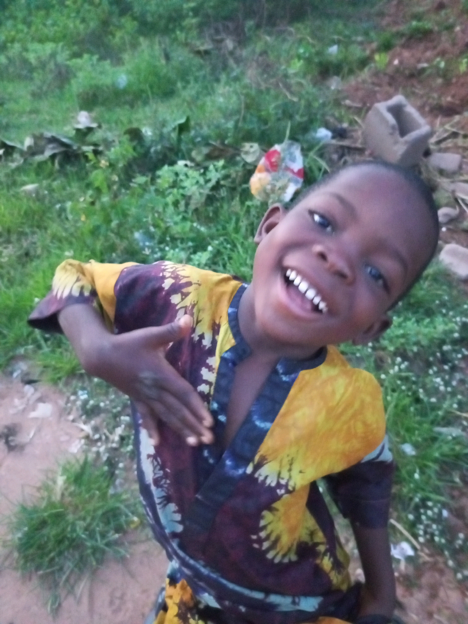 A young Black child, appearing to be a boy, smiles broadly at the camera. He is wearing a vibrantly colored shirt with a tie-dye pattern of yellow, purple, and blue. His left hand is raised, fingers spread in a gesture. The child's eyes are dark, and his teeth are visible in his wide smile. The background is out of focus, showing a mixture of green vegetation, dirt, and what appears to be some discarded debris. The lighting suggests it is daytime, possibly late afternoon due to the muted tones. The environment appears to be outdoors, in a non-urban setting. The location is unknown, but the question mentions Tempure, Nigeria.