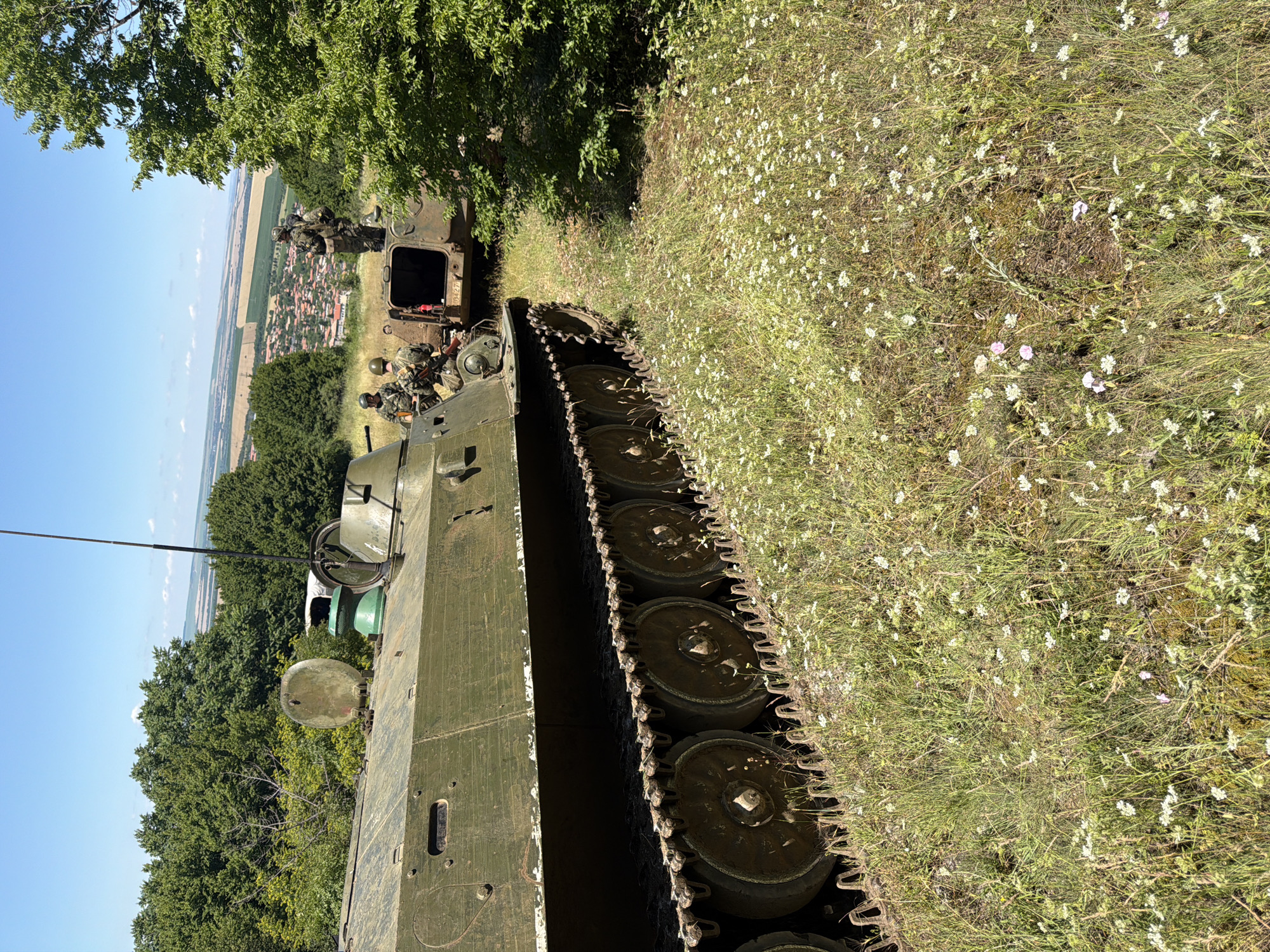 In Samovodene, Bulgaria, a military training exercise is underway. Two tracked military vehicles are positioned on a grassy hillside dotted with small white flowers. The foreground vehicle is a green armored personnel carrier, its tracks and road wheels visible, positioned at an angle to the viewer. Behind it, a second, similar vehicle, also green, is open at the rear. Several soldiers in military attire are positioned on and around the vehicles. One soldier stands on the rear deck of the second vehicle, looking out over a valley. Another soldier is seated on the edge of the first vehicle. The background reveals a clear blue sky, a line of dense green trees, and a small village with red-tiled roofs in the valley below, extending towards agricultural fields and a distant horizon. The overall scene depicts a military activity in a rural, natural environment.