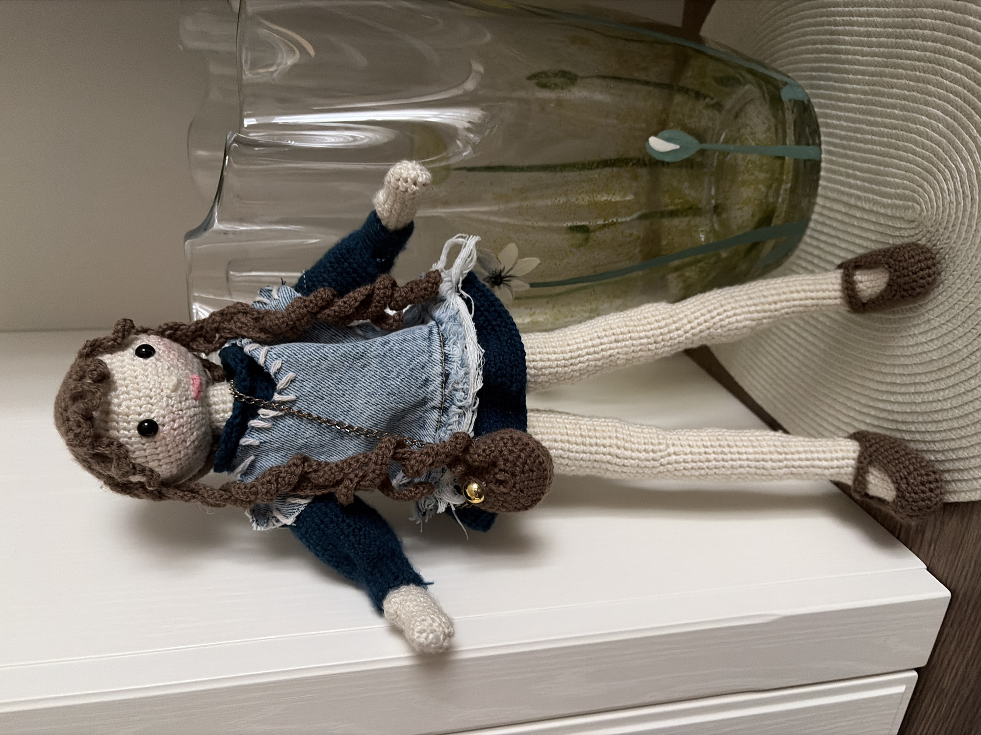 A handmade crochet doll stands upright on a round, textured mat. The doll has light-colored skin, brown yarn hair styled in braids, and black button eyes. It wears a dark blue knitted long-sleeved shirt, a denim dress with a white frayed hem, and dark blue knitted leggings. Brown crochet shoes adorn its feet, and a small brown crochet purse with a gold charm hangs from a silver chain across its chest. The doll is positioned next to a large, clear glass vase decorated with a green floral pattern. In the background, a white door frame is visible on the left, and a wall appears on the right. This scene does not provide any information to suggest it is located in Astana, Kazakhstan.