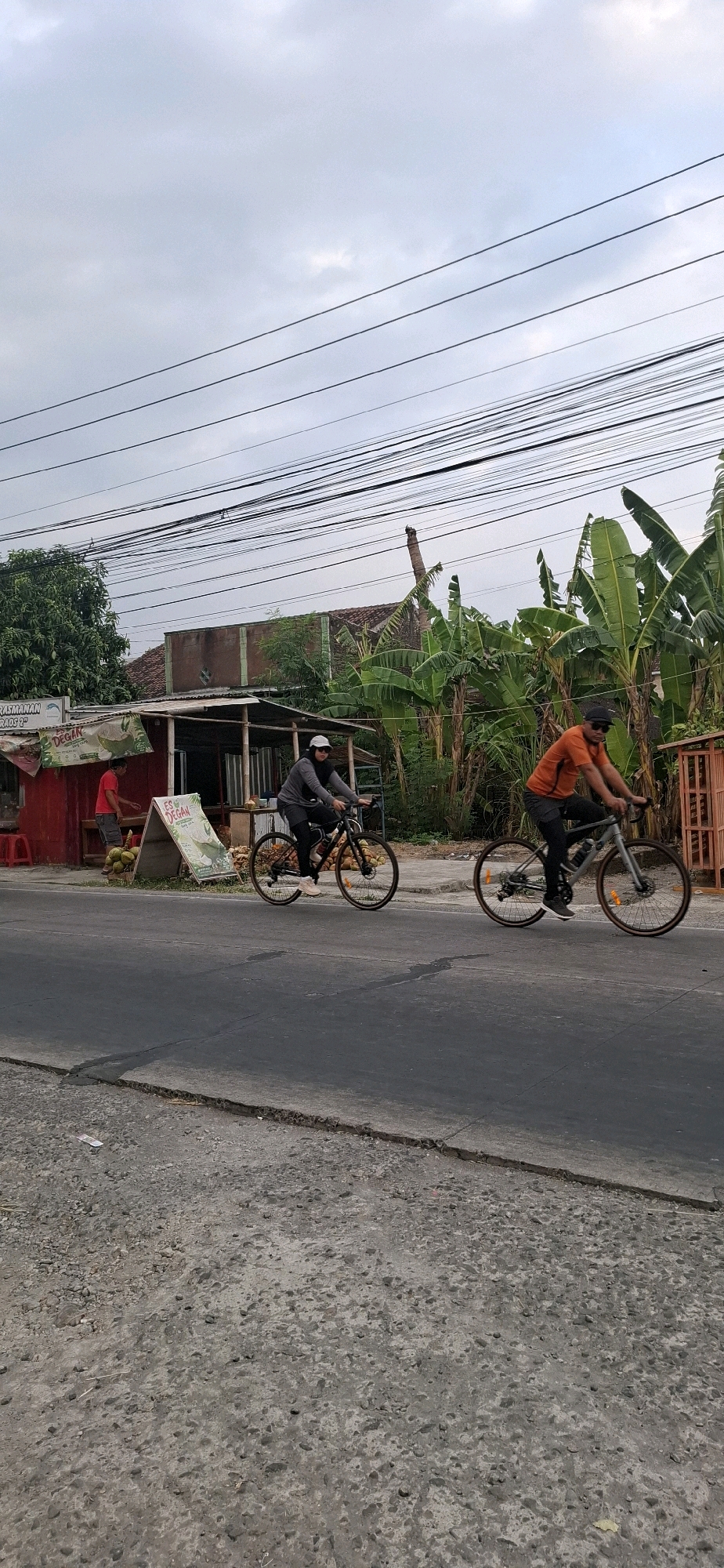 The image depicts an outdoor scene in Desa Dibal, Indonesia, under a cloudy sky. The foreground shows a paved road with some gravel on the shoulder. In the middle ground, two individuals are riding bicycles on the road. The person on the left is wearing a grey long-sleeved shirt, black leggings, and a white headscarf, riding a dark-colored bicycle. The person on the right is wearing an orange t-shirt, black pants, and a black baseball cap, riding a light-colored bicycle.
Behind the road, there is a small roadside establishment with a red facade and a corrugated metal roof, selling what appear to be coconuts and drinks. A person in a red t-shirt is standing near the establishment. To the right of the establishment, a dense cluster of banana trees is visible, with some houses and structures further in the background. Overhead, numerous electrical wires are strung across the frame. The overall atmosphere is that of a typical rural or semi-urban roadside scene in Indonesia.