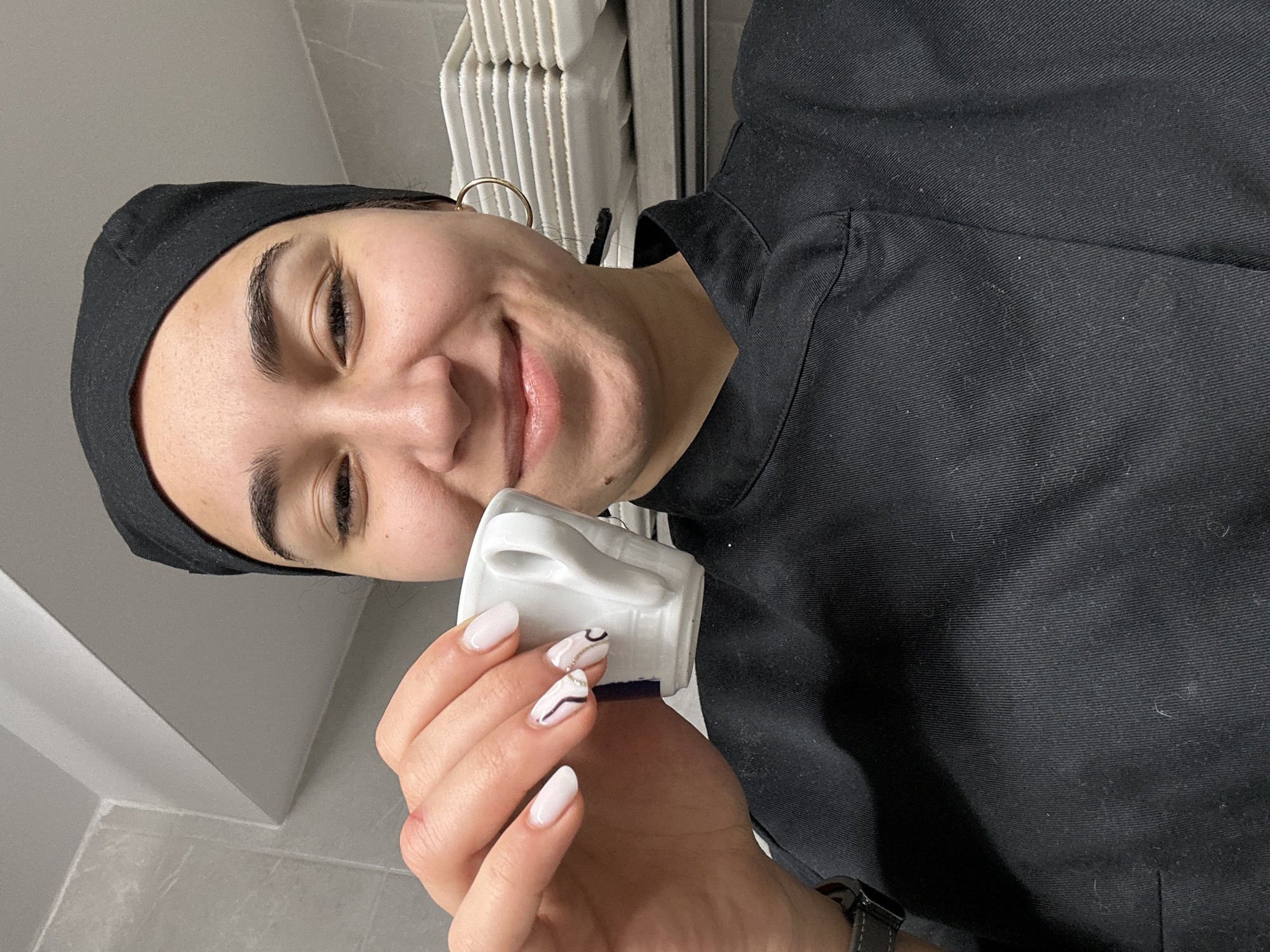 A person wearing a black surgical cap and a black shirt is holding a white mug, close to their face. The person is smiling and wearing a gold hoop earring. In the background are white objects stacked on a shelf, and a gray wall. The person has a watch on their wrist, and painted fingernails. The city of Burgas, Bulgaria, is not discernible.