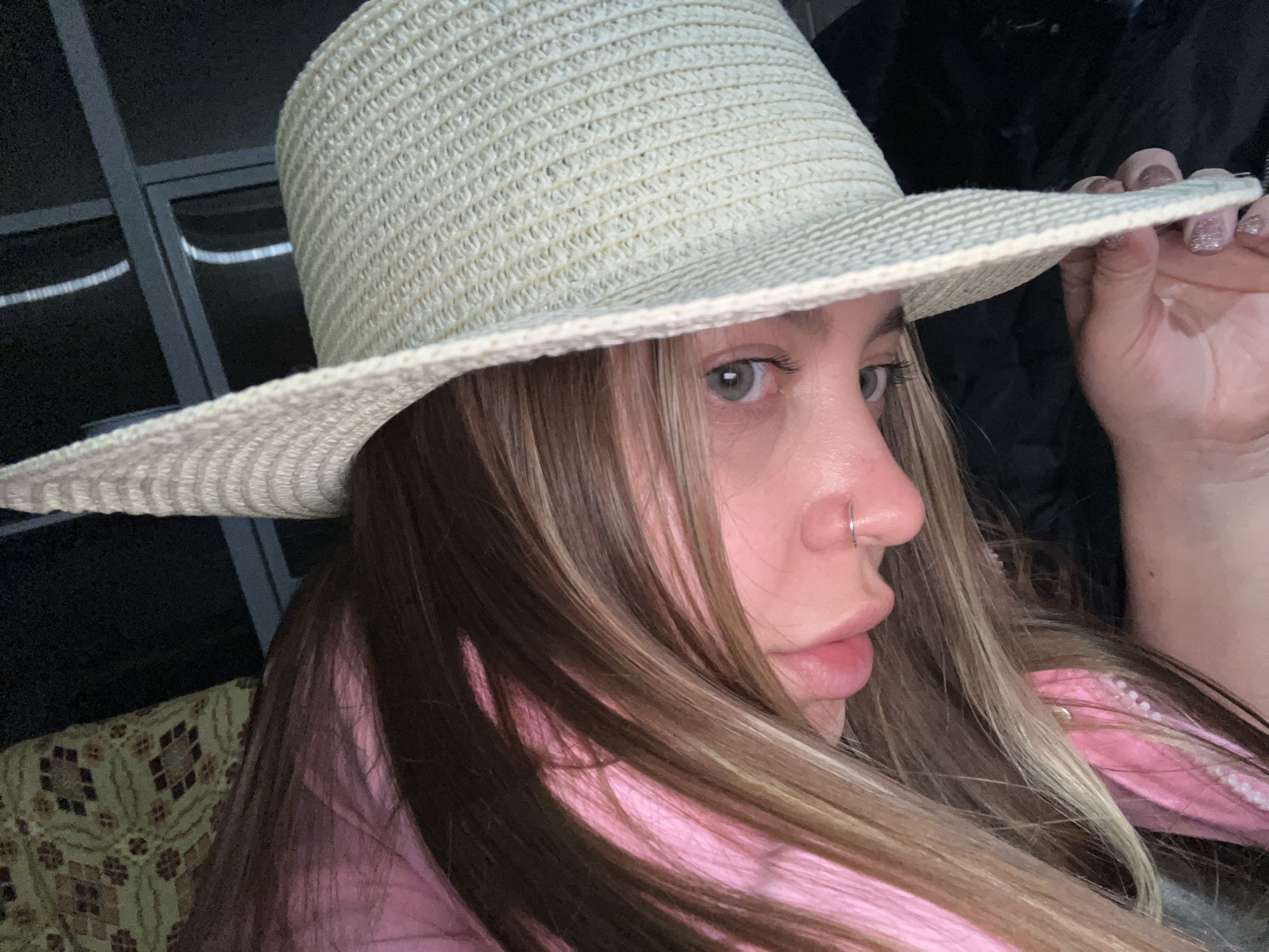 A woman with long blonde and brown hair wearing a pink shirt and a light-colored straw hat is looking off to the side. She has a nose ring. Her hand is holding the brim of the hat. A patterned object is visible behind her. The location is presumed to be Novopokrovskaya, Russia.