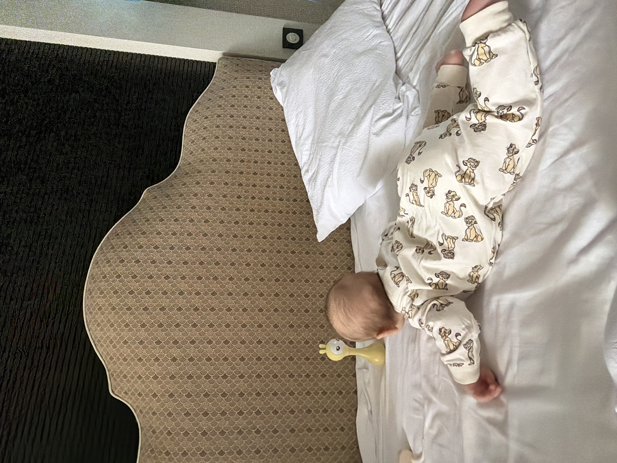 The image shows a baby lying on a white bed with a white pillow, next to a beige headboard with a scale pattern and a dark brown wall. The baby is wearing a white onesie with a "Lion King" print. A yellow giraffe toy is near the baby's head. The location is Khimki, Russia.