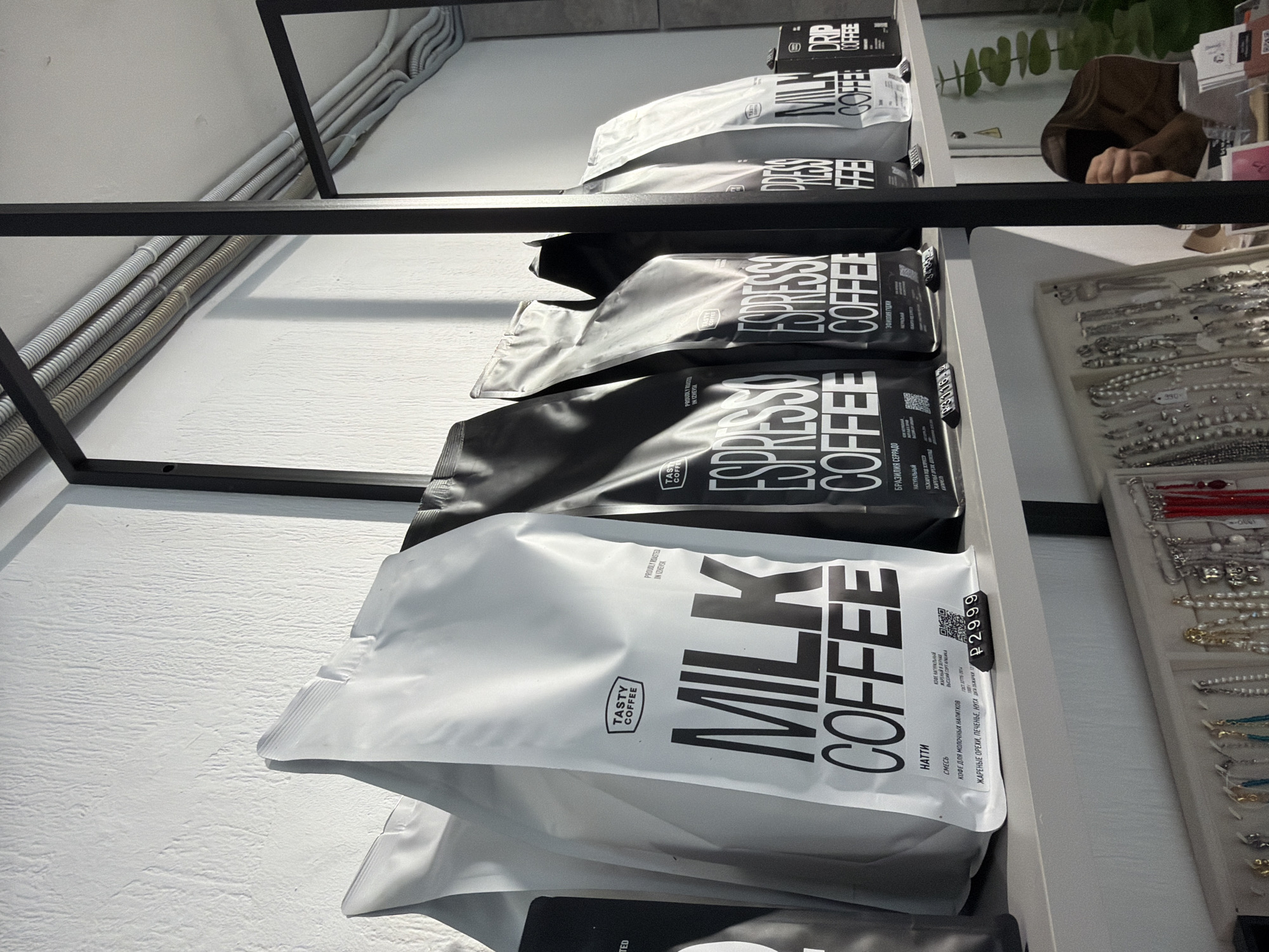 The image displays an interior retail setting in Chaikovsky, Russia, showcasing products on a light gray shelf supported by a black metal frame. In the foreground, several large stand-up pouches of "Tasty Coffee" are prominently displayed. Two white pouches, labeled "MILK COFFEE," are visible on the left, one bearing a price tag of "P 2999" and a QR code. Adjacent to these are multiple black pouches, labeled "ESPRESSO COFFEE," also with price tags (e.g., "P30118"). Some packaging indicates "PROUDLY ROASTED IN IZHEVSK." A small black box labeled "DRIP COFFEE" is visible on the far right of the shelf.
Below the main shelf, a lower display case exhibits various jewelry items, including necklaces and bracelets, on white stands. In the bottom right corner, the upper back of a person with brown hair, wearing a brown garment, is partially visible, appearing to be bent over or attending to something on a lower counter. Green foliage from a plant is visible behind this individual. The background consists of a textured white wall with exposed corrugated electrical conduits running horizontally near the ceiling.