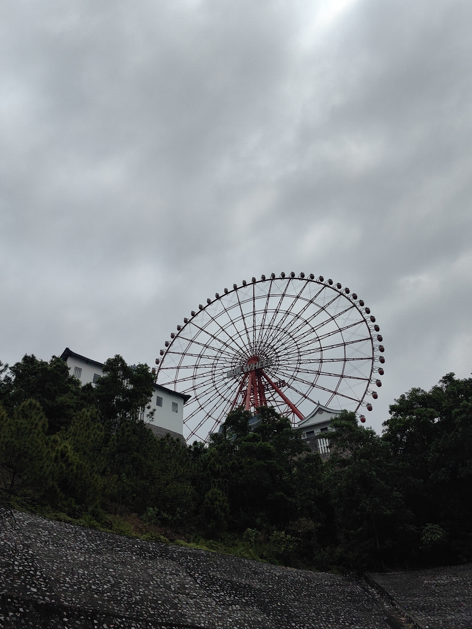 The image depicts a large Ferris wheel partially obscured by trees and a building on a hill in Hong Gai Ward, Vietnam. The sky is overcast with grey clouds. The Ferris wheel has a red frame and support structure with passenger cars around its circumference. The building is white with a dark roof, visible through the trees. A stone structure slopes downwards in the foreground.