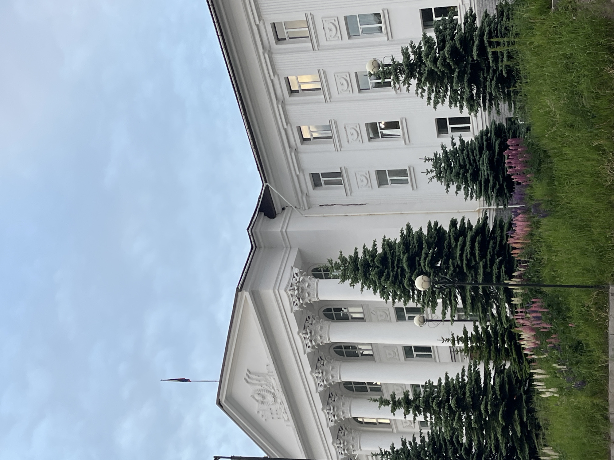 A low-angle, outdoor, medium shot shows the white facade of a large, classical-style building in Petropavlovsk-Kamchatsky, Russia. The building features a prominent pediment with relief carvings and fluted Ionic columns supporting a triangular roof. Multiple windows line the building's facade, with some reflecting the cloudy sky. Several evergreen trees are planted in front of the building, and a bed of lupine flowers in shades of pink, purple, and white blooms in the foreground. Two ornate lampposts stand amongst the trees and flowers. A flag flies from a flagpole on the roof of the building. The sky is overcast and a pale blue.
