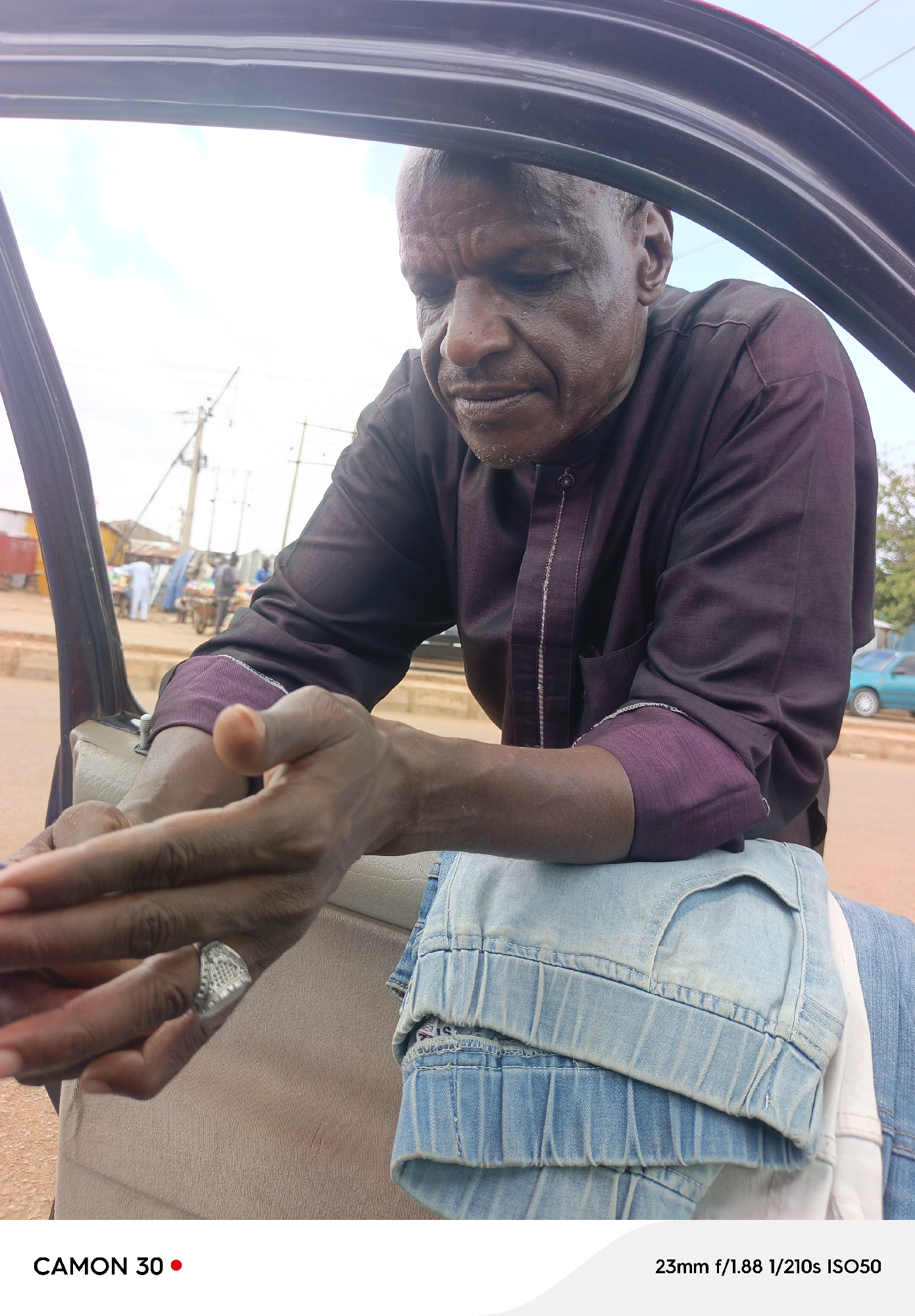 A man is pictured in a close-up, seated in the open door of a vehicle. He is wearing a dark purple, collared shirt with a vertical white stripe down the front and embroidered trim on the cuffs. His hands are resting on his lap, with a silver ring on his left ring finger. He is looking downwards, with a pensive expression. His jeans are folded and resting on his lap, a light blue denim material.
In the background, beyond the man and the open car door, a street scene is visible. There are buildings, utility poles, and some people are walking in the distance. A blue car is parked on the side of the road. The lighting suggests it is daytime, with bright sunlight. The overall atmosphere is casual and possibly a moment of quiet reflection for the man. The text "CAMON 30" is visible in the bottom left corner, and "23mm f/1.88 1/210s ISO50" is visible in the bottom right corner, indicating the camera settings used for the photograph.