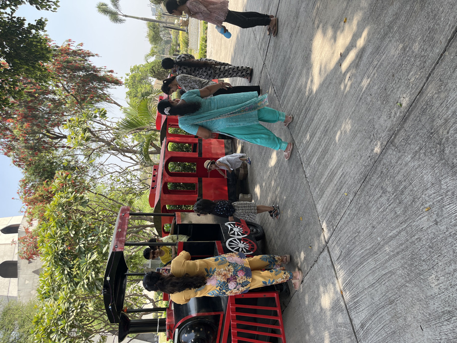 The image shows a group of people standing around a toy train in a park. The train is red and black and has a number of carriages. The train is parked on a paved area. There is a woman in a yellow and floral dress standing next to the train and looking at a boy inside the train. Another woman in a light blue dress is standing next to the train and looking at the boy. There are two girls in front of the train, a woman and a girl standing next to the train, and a woman in black standing behind the train. The women are dressed in traditional Indian clothing. The park is well-kept and has trees and grass. This image is in Sahibzada Ajit Singh Nagar, India, at the coordinates 30.703, 76.668, which correspond to a public park called the 'Park of Sahibzada Ajit Singh Nagar'. The image was taken on a sunny day. The shadows suggest it is mid-morning or late afternoon.
