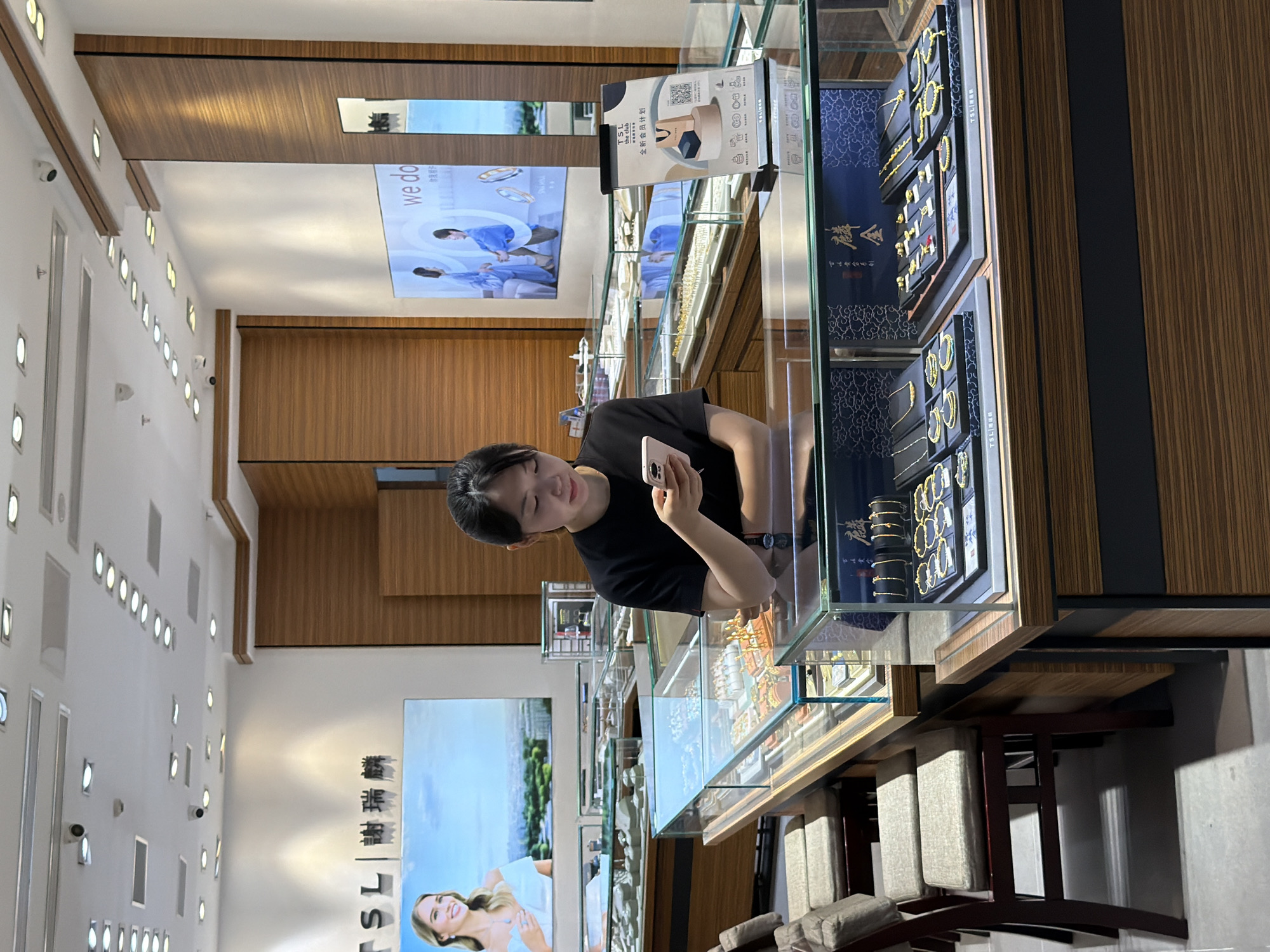 In a well-lit jewelry store, a young woman with black hair, wearing a black t-shirt, stands behind a glass display counter. She is looking down at her mobile phone, holding it in her hands. The display cases in front of her showcase a variety of gold necklaces and bracelets. Behind her, large screens display advertisements for jewelry. The store features polished wood paneling on the walls and ceiling. To the left, a row of chairs with padded seats are arranged along another display counter. The overall environment suggests a modern and upscale retail setting. The location is Yuzhong District, China.