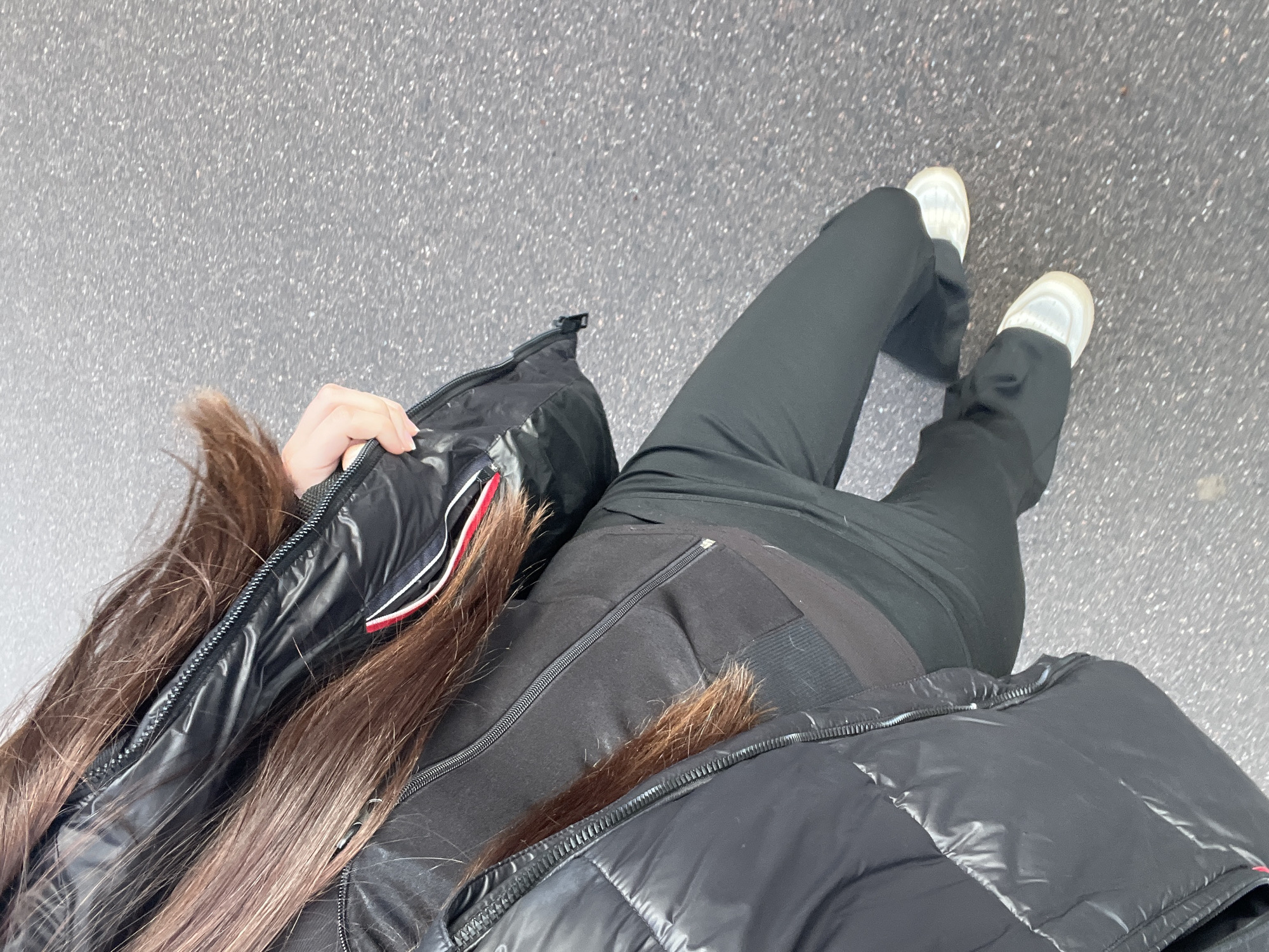A person wearing black clothing, including a glossy puffer jacket, black pants, and white sneakers, stands on a gray asphalt surface. Part of their face is obscured by the jacket and long brown hair. The perspective is from above, looking down. The location is unidentifiable as Saint Petersburg, Russia.