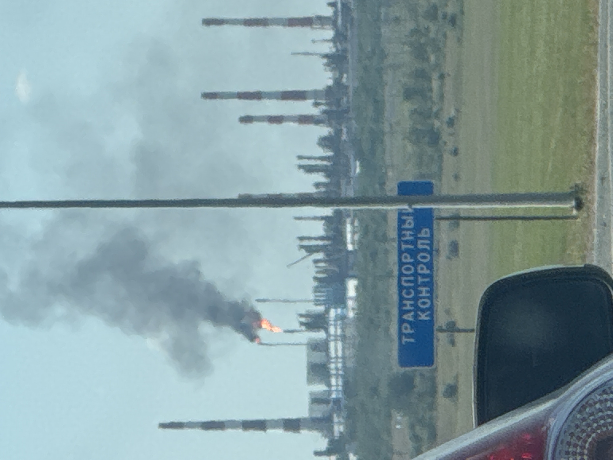 A plume of black smoke and orange flames erupts from an industrial facility. The facility is characterized by a cluster of tall, striped smokestacks and other industrial structures. In the foreground, a blue sign with white Cyrillic text, which reads "ТРАНСПОРТНЫЙ КОНТРОЛЬ" (TRANSPORT CONTROL), is visible, mounted on a metal pole. Part of a vehicle, including a side mirror and taillight, is present in the lower left corner of the frame. The background consists of a hazy sky and a landscape with scattered vegetation. The scene appears to be in the city of Kholodnye Kliuchi, Russia, given the context of the sign.