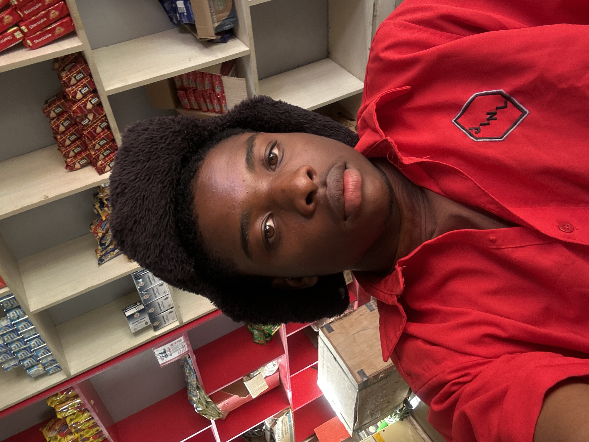An eye-level portrait features a young, dark-skinned individual with brown eyes and a neutral expression, looking directly at the camera. They wear a dark, fluffy hat or wig and a bright red collared shirt. The shirt bears a hexagonal logo with a jagged line and the letters "JINL" (or "SINL") embroidered in white and black.
The person is positioned indoors, with a background of shelves filled with various packaged goods. The shelves are a mix of light beige and vibrant red sections. On the shelves, numerous red packages of "Shortcake" biscuits are visible, stacked neatly. Other shelves hold blue and silver packages, partially identifying "Golden Penny" products, and some yellow-orange packaged items. Several cardboard boxes are also present, some appearing empty or partially used.
The overall setting suggests a small retail store, convenience shop, or a storage area within a commercial establishment in Jos, Nigeria. The lighting is artificial, illuminating the scene. The image is oriented sideways, making the shelves and person appear tilted. No specific activity is depicted other than posing for the photograph.