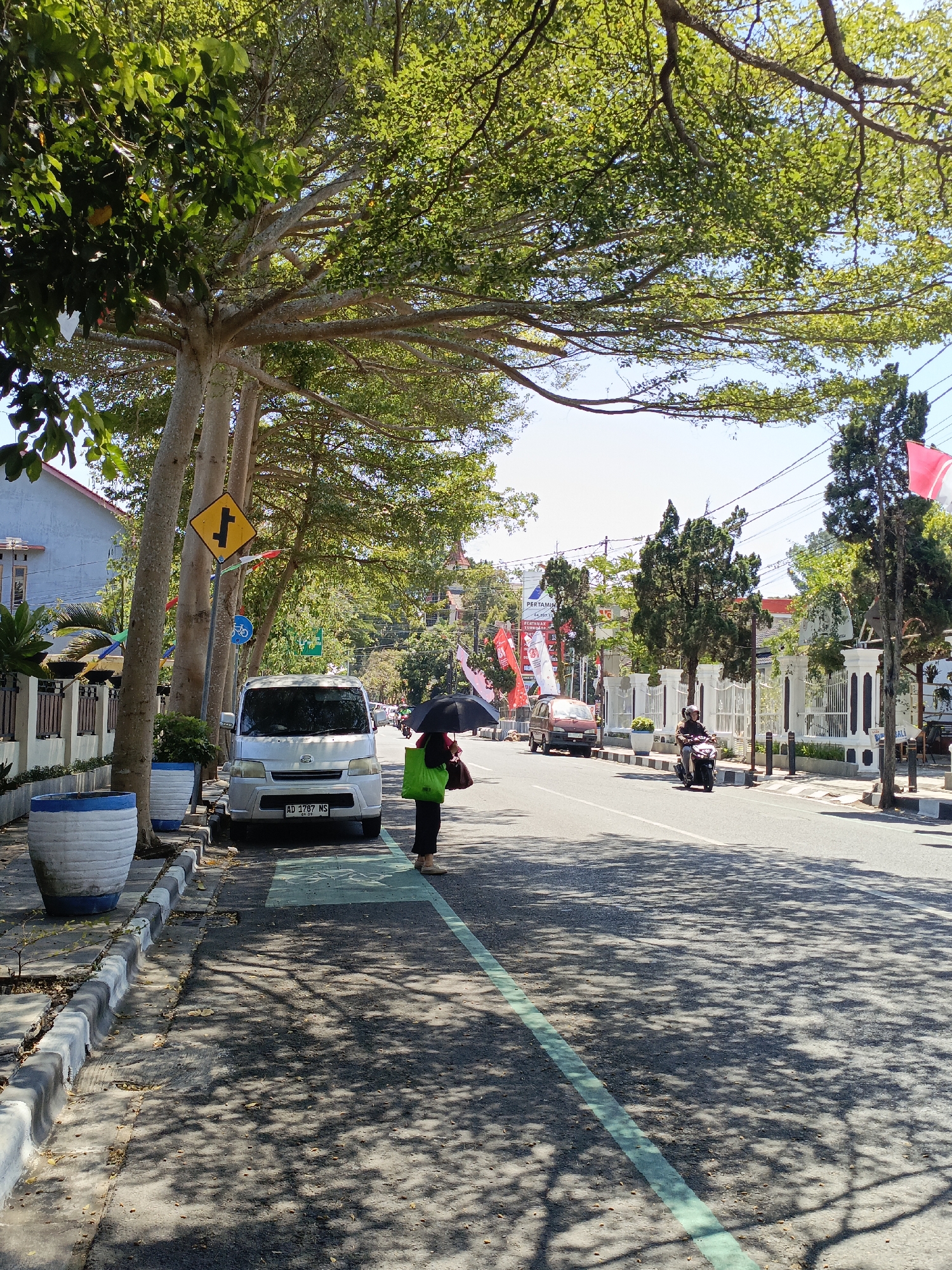 A street scene in Salatiga, Indonesia, captures a person holding a black umbrella and a green bag, walking on the left side of the road, partially shaded by large trees. The person is wearing dark clothing. A white minivan with license plate "AD 1787 NS" is parked on the side of the road. The street is paved, with a green painted lane marker. Various trees line the sidewalk, casting dappled shadows on the ground. Buildings and white decorative pillars are visible in the background. Several red and white flags are displayed. A motorcycle is being ridden on the right side of the road. The sky is clear and blue.