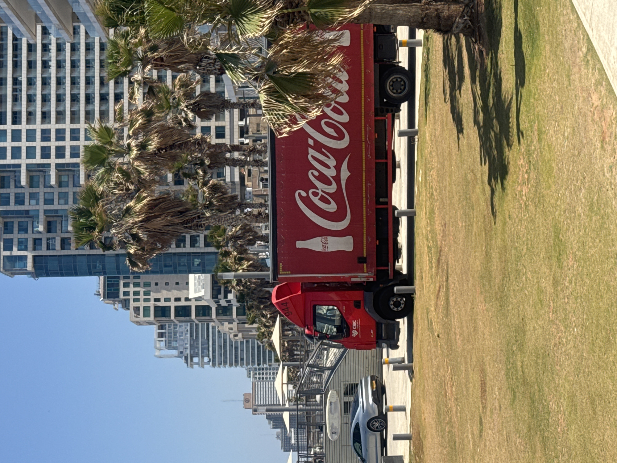 A Coca-Cola truck parked on a street in Tel-Aviv, Israel, on a clear, sunny day. The street runs along a grassy area, with palm trees casting shadows onto the grass. Modern high-rise buildings line the opposite side of the street, forming a city skyline. A silver car is parked behind the truck. A pedestrian walkway with barriers is present between the street and the grass.