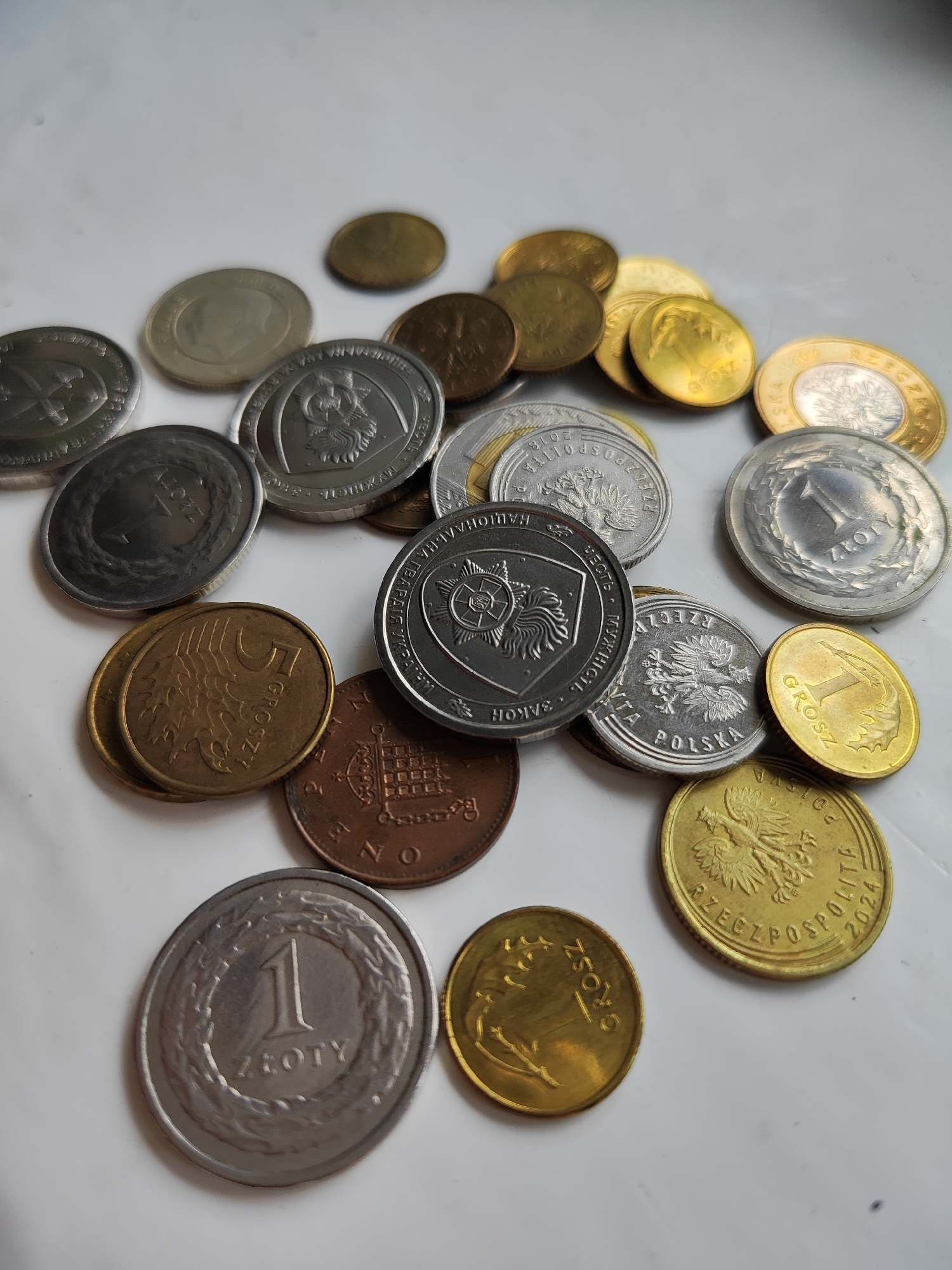 The image displays a collection of assorted coins scattered on a white, textured surface. The coins are of various sizes, colors, and designs, suggesting they are from different countries and denominations. Some coins are silver-toned, while others are golden or coppery. Several coins feature intricate designs, including eagles, portraits, and inscriptions in what appears to be a Slavic language. The lighting suggests an indoor environment, possibly a table or countertop. There is no indication of the city of Korosten, Ukraine, in the image. The scene is static, with no actions or interactions taking place.
