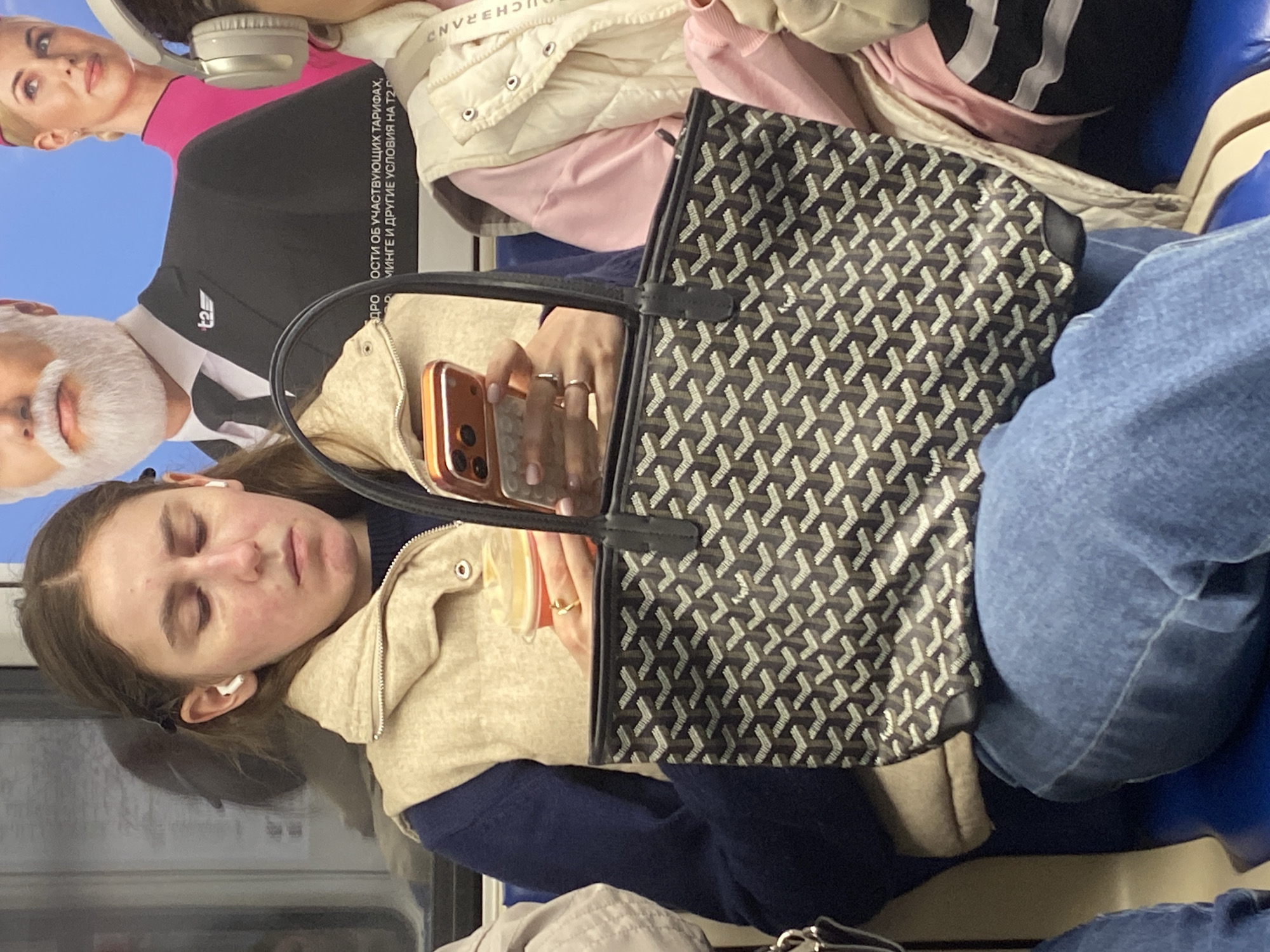 A woman with closed eyes and earbuds is seated on public transport, possibly a train or bus, with a tan jacket, blue sweater, and blue jeans. She's holding a smartphone and a geometric-patterned handbag rests on her lap. Behind her, an advertisement features a man and woman in formal attire, overlaid with Russian text. The specific location in Khimki, Russia is impossible to determine from the image alone.
