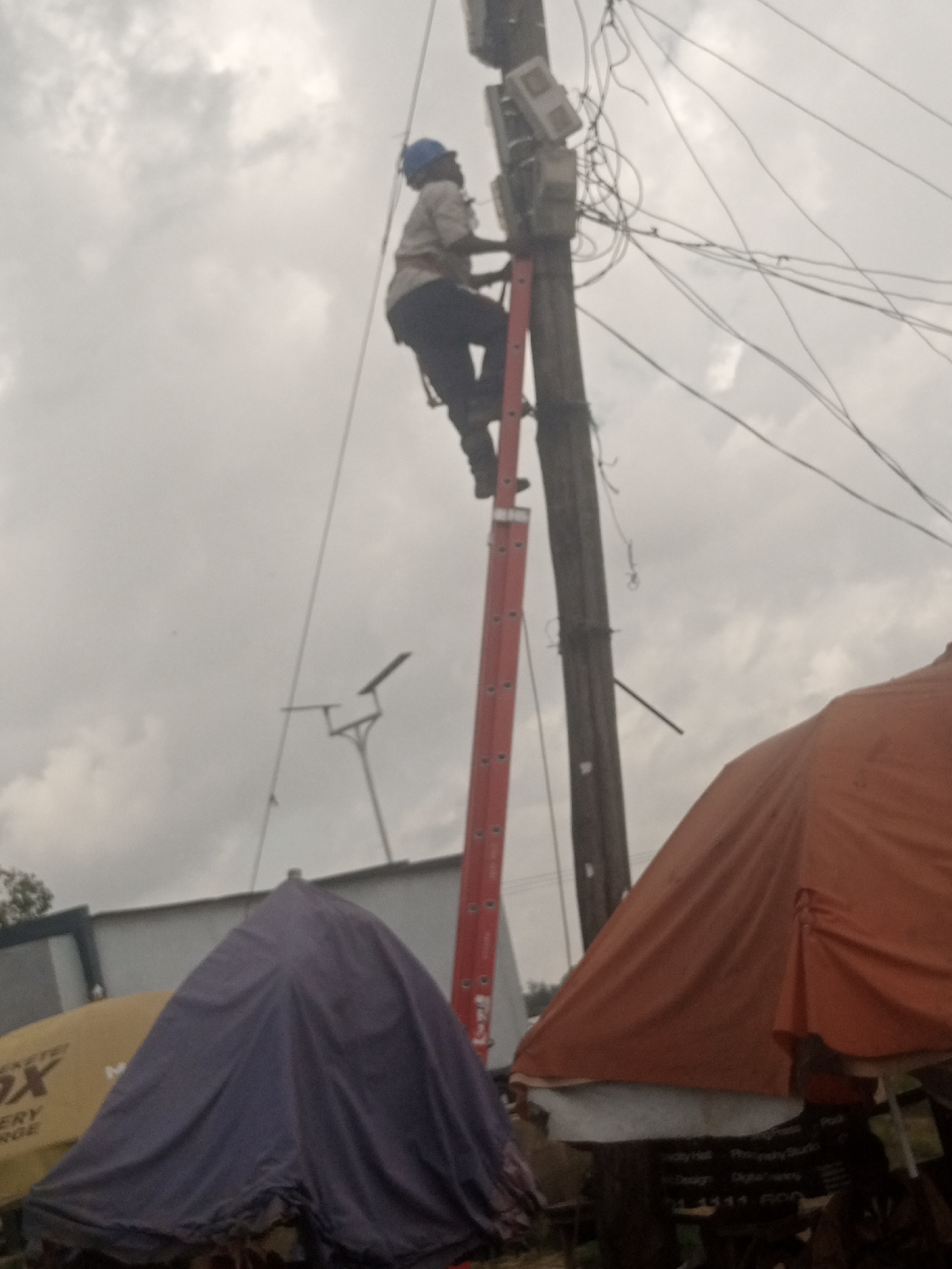 A person wearing a hard hat and work clothes is on a tall ladder, working on a utility pole with many wires and electrical meters. The person is positioned near the top of the ladder, holding onto the pole. In the foreground, there are several market stalls covered with tarps, one purple and one orange, suggesting an outdoor market setting. The sky is overcast. The scene appears to be in an urban environment, though the specific city and location within it are unknown.