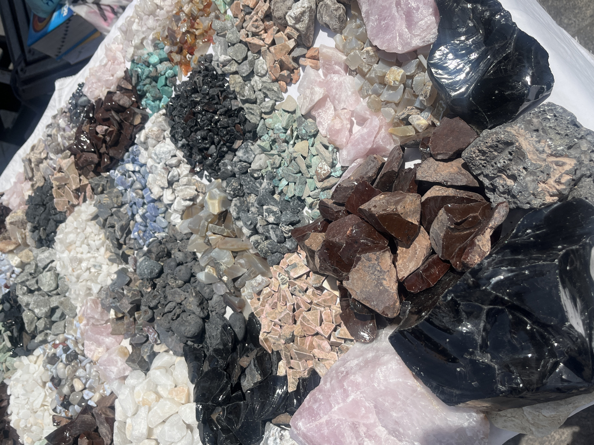 The image shows a close-up, eye-level view of a variety of rocks and minerals displayed on a white surface, possibly a cloth or table. The scene is in a city named Unknown, Unknown. The collection includes a range of colors and textures, from smaller, granular stones to larger, more polished pieces. Notable materials include rose quartz (pink), obsidian (black and shiny), and various shades of gray, brown, and blue-toned stones. The rocks are arranged in distinct groupings, possibly for sale or display. The lighting suggests an outdoor setting, likely during daylight hours. There are no people directly visible in the frame.