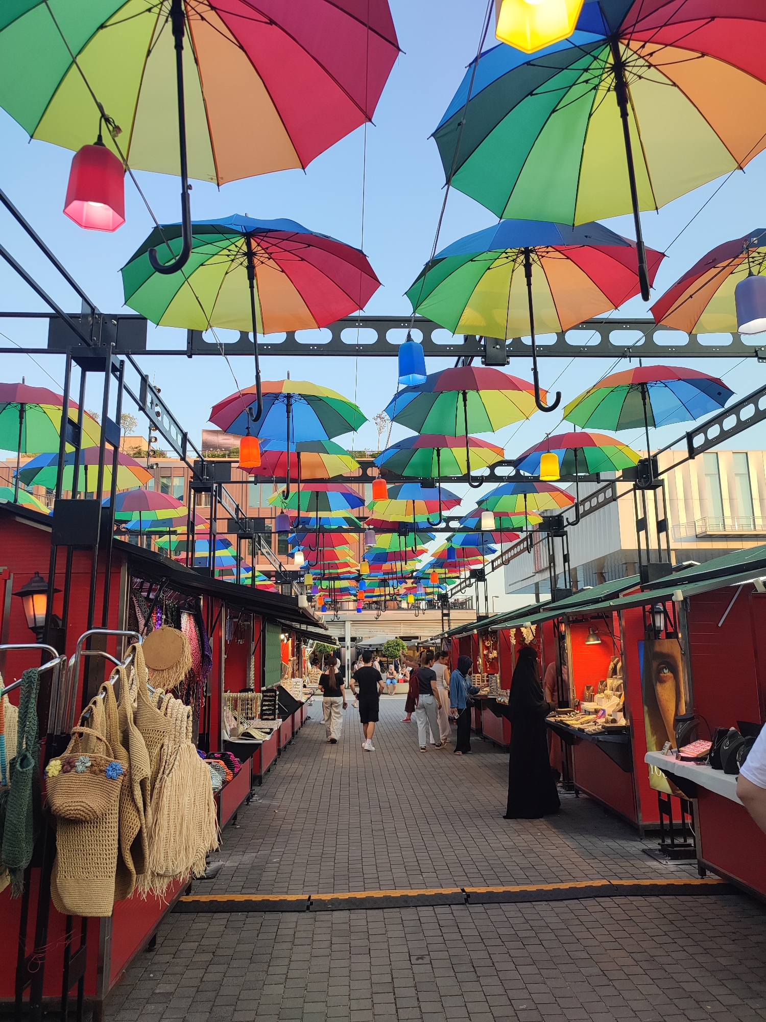 This image captures a vibrant street market scene in Beyoğlu, Turkey, characterized by an overhead canopy of colorful, rainbow-hued umbrellas suspended against a clear blue sky. The umbrellas are arranged in rows, creating a visually striking pathway. Below the umbrellas, the market is lined with red stalls, some displaying merchandise like woven bags and jewelry. Several people are seen walking along the cobblestone pathway and browsing at the stalls. The overall atmosphere suggests a lively outdoor market, possibly in a pedestrianized area of the city.
