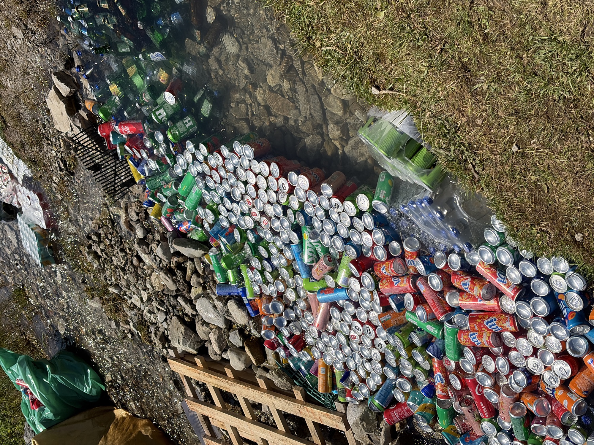 In Gusinje, Montenegro, a shallow, rocky stream is heavily polluted with discarded aluminum cans and plastic bottles. The debris is concentrated in the water and along the banks. Among the litter are green and red aluminum cans, some appearing to be soda or beer cans, and clear plastic bottles, some containing blue liquid. In the upper left corner, a large, crumpled green plastic bag is visible, partially obscuring a brown bag. Wooden pallets are stacked on the left bank, and a black plastic grate is submerged in the water near the debris. The surrounding environment consists of grass and rocks.