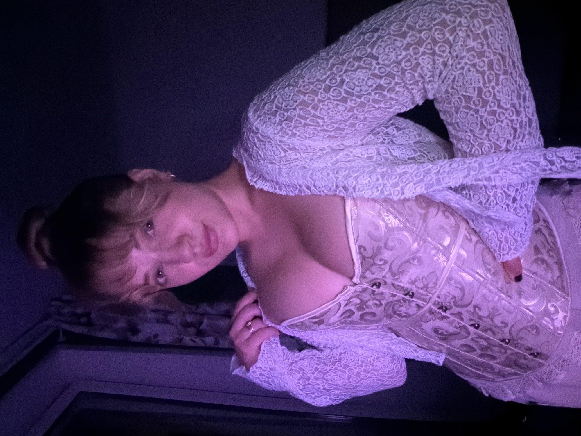 This image, taken in Большая Шильна, Russia, captures a person, likely a young woman, in an intimate indoor setting. She is positioned with her head tilted to her left, looking directly at the camera. The scene is dimly lit with strong ambient purple and pink lighting, casting a soft glow on her face and upper body against a dark, featureless background.
She is wearing a two-piece ensemble. An outer layer consists of a white, long-sleeved garment made of delicate lace, featuring an intricate, repeating circular or floral pattern. Beneath this, she wears a light-colored, possibly cream or pale gold, corset-style top. This inner garment has a subtle, textured pattern, resembling damask or scrollwork, and visible vertical boning. Small, dark circular embellishments, possibly studs or eyelets, are visible along its structure.
Her hair appears light-colored, perhaps blonde or light brown, styled with bangs across her forehead. A ring is visible on the fourth finger of her left hand, and her fingernails are painted with dark polish. The surface her head rests on appears to be covered with a subtly patterned fabric. The overall mood is atmospheric and suggestive, created by the distinct lighting and direct gaze. No text is visible in the image.