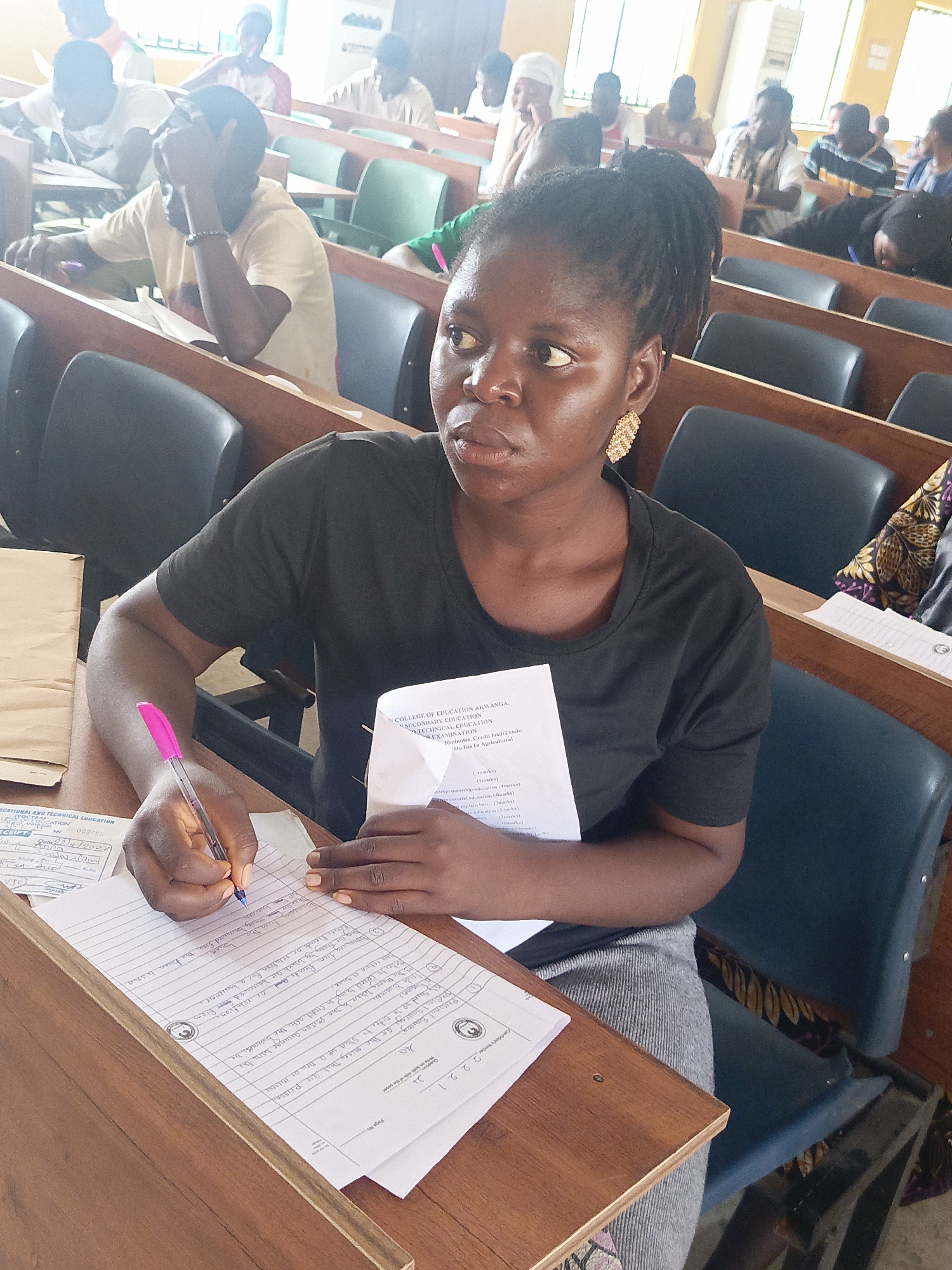 In a classroom in Akwanga, Nigeria, a young woman with her hair pulled back is seated at a desk, writing on a paper with a pink pen. She wears a black shirt and has on gold earrings. A test paper from the "College of Education Akwanga" lies in her other hand. The classroom is filled with other people, some writing, and others looking around. The chairs are a mix of green and dark blue. The desks are wooden, and the room appears to be well-lit, likely with natural light.