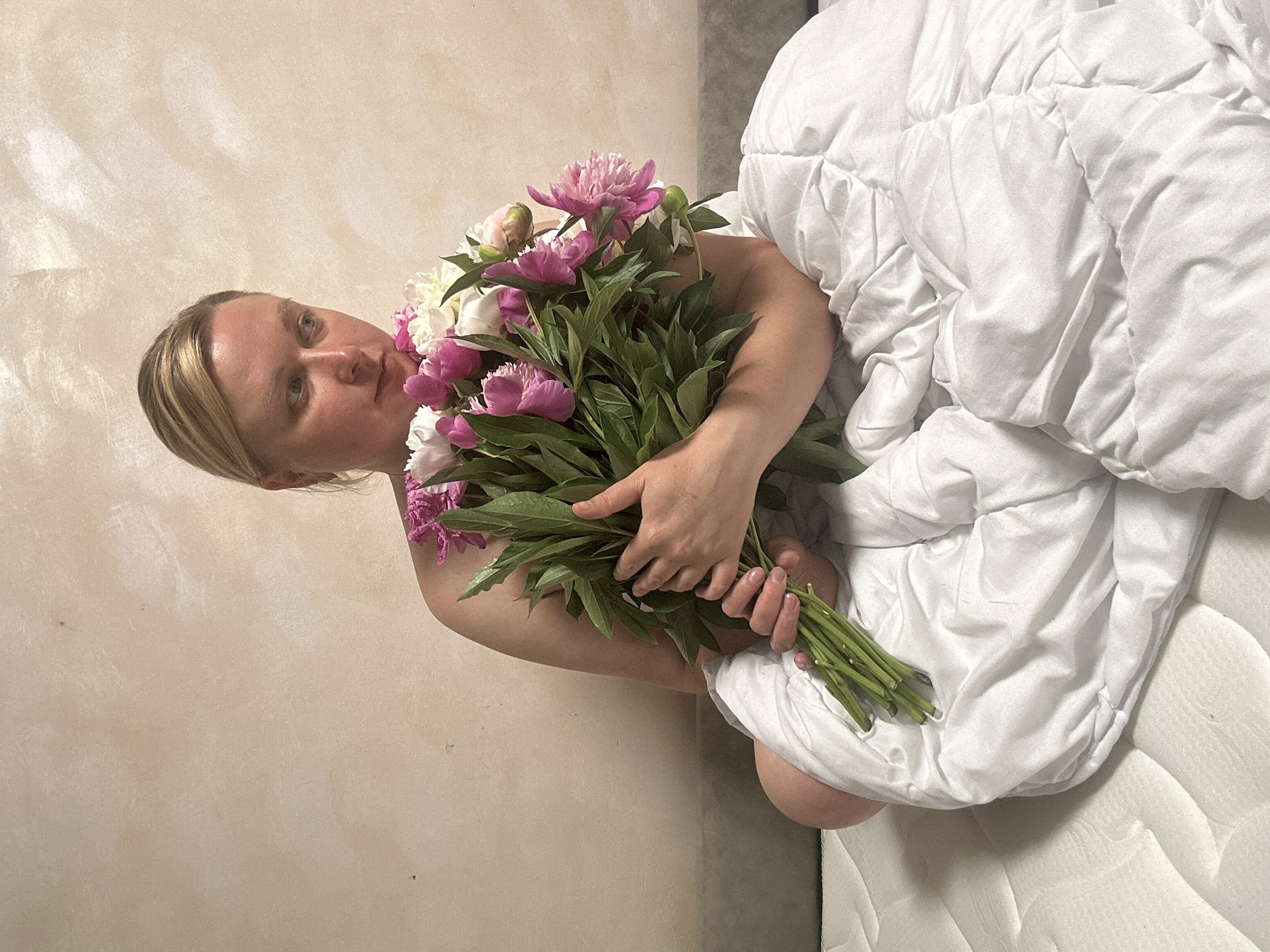 A woman is sitting on a mattress, wrapped in a white duvet, and holding a bouquet of pink and white peonies. She has blonde hair pulled back and is looking down at the flowers. The background is a light-colored textured wall. This image was taken in Chaikovsky, Russia.
