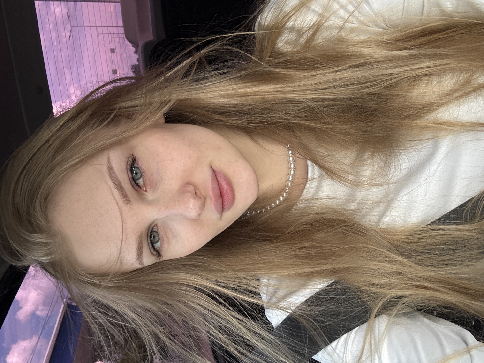 The image captures a young woman in a close-up, diagonally oriented portrait, likely taken inside a vehicle. She has long, light blonde hair that cascades around her shoulders and chest. Her striking green eyes gaze directly towards the viewer. Her fair skin shows subtle freckles across her nose and cheeks, and her lips are naturally pink and slightly parted. She is wearing a plain white top and a delicate pearl necklace. A dark seatbelt crosses her chest, reinforcing the setting within a car.
Through the car window in the background, a sky with prominent purple and pink hues is visible, suggesting either sunrise, sunset, or the effect of a tinted window. Faint clouds and dark, indistinct structures resembling power lines or distant vegetation are silhouetted against the colorful sky. The woman's expression is calm and direct. No activity beyond posing for the photograph is depicted. No visible text is present in the image.