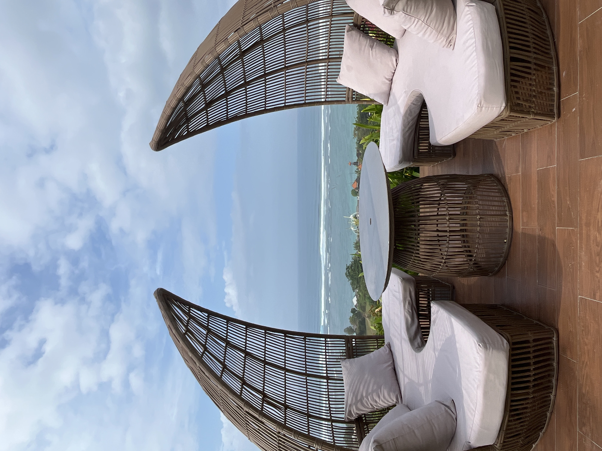 An outdoor seating area offers a panoramic view of the ocean with a cloudy sky above. The seating arrangement consists of two curved, wicker-style daybeds facing each other, with light-colored cushions and pillows. A round wicker table with a dark tabletop is positioned between the daybeds. The furniture is placed on a wooden deck, with lush greenery visible in the background leading down to the coastline. The ocean exhibits gentle waves breaking on a distant shore. In the far distance, some buildings and a tower are visible. The scene appears to be from a resort or restaurant in Kuta Selatan, Indonesia, likely on a cliff or elevated location overlooking the sea. There are no people present in the image.