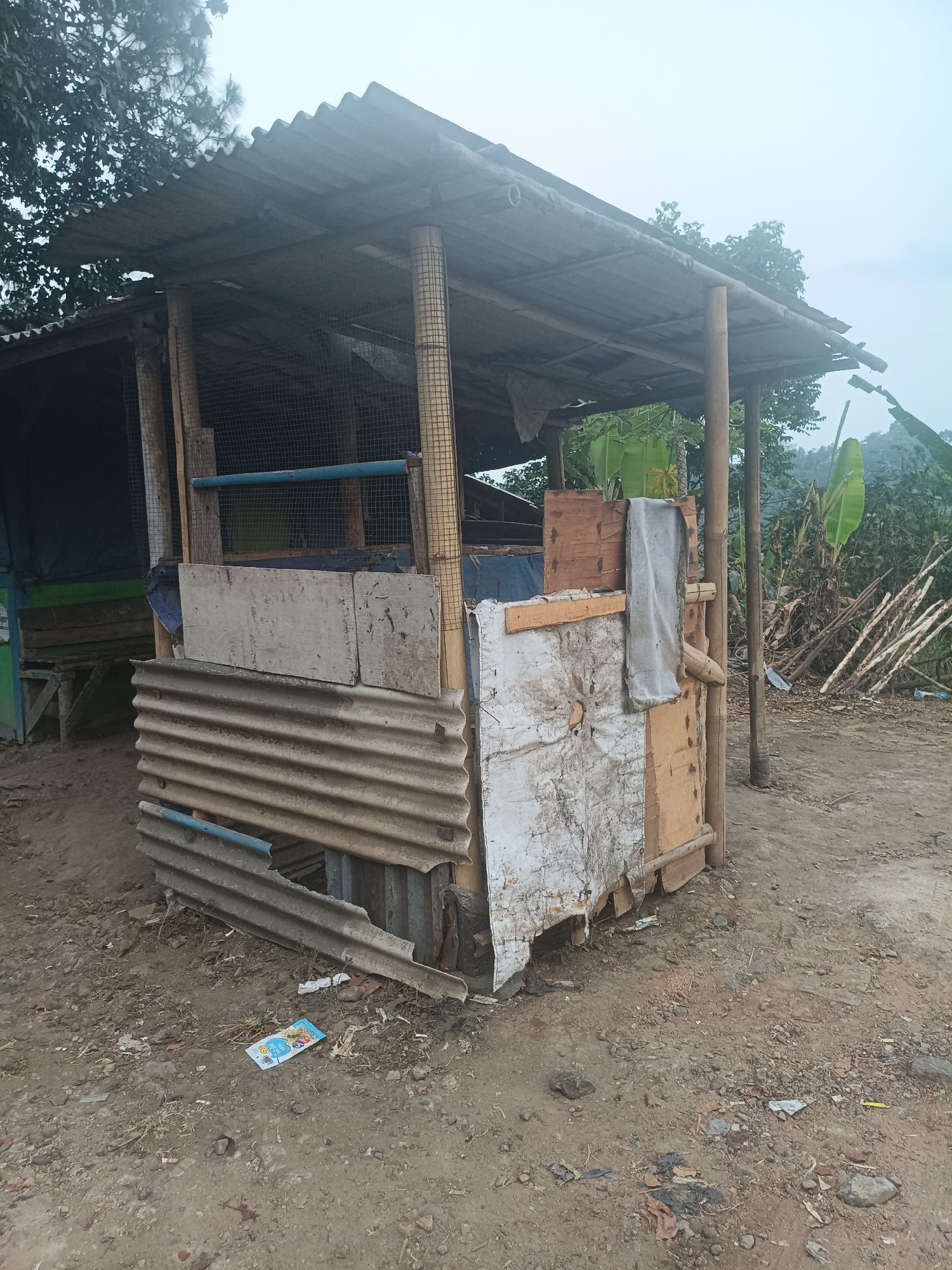 In Kadununggal, Indonesia, a small, makeshift structure stands on uneven, dusty ground. The structure appears to be a shelter or stall, constructed from salvaged materials including corrugated metal, bamboo poles, wood planks, and wire mesh. The roof is made of corrugated metal and is supported by bamboo posts. The walls are a patchwork of different materials, with some sections covered in distressed white material, others with wood, and a lower section made of corrugated metal sheets. A blue pipe runs horizontally along the upper portion of the structure, and a gray tarp-like material is draped over a bamboo support on one side. In front of the structure, on the ground, are scattered debris, including small stones, dry leaves, and a piece of colorful packaging. Behind the structure, lush green vegetation, including banana trees, is visible, with a hillside in the distance. The overall impression is one of a rural or semi-rural environment, with evidence of resourcefulness in its construction.
