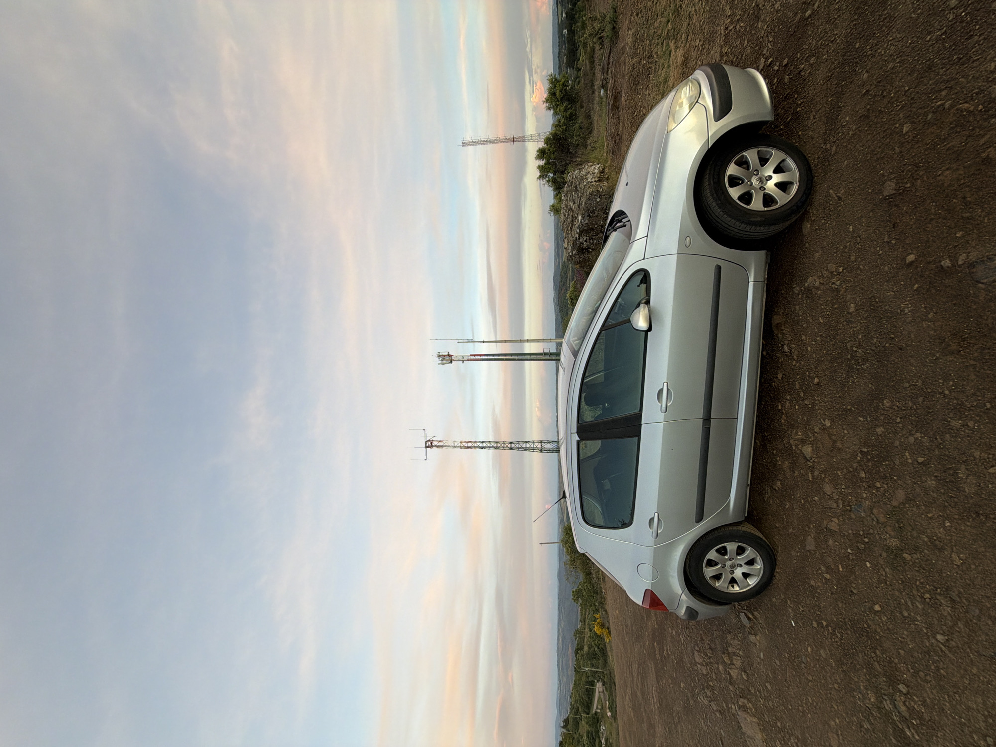 The image shows a silver Peugeot 307 hatchback parked on a dirt track in a rural area in Bragança, Portugal. The car is facing toward a sky filled with pastel-colored sunset clouds. Three cell towers can be seen in the background, in the center of the image. The ground is brown and rocky. There are green trees and vegetation in the distance. No people are visible.