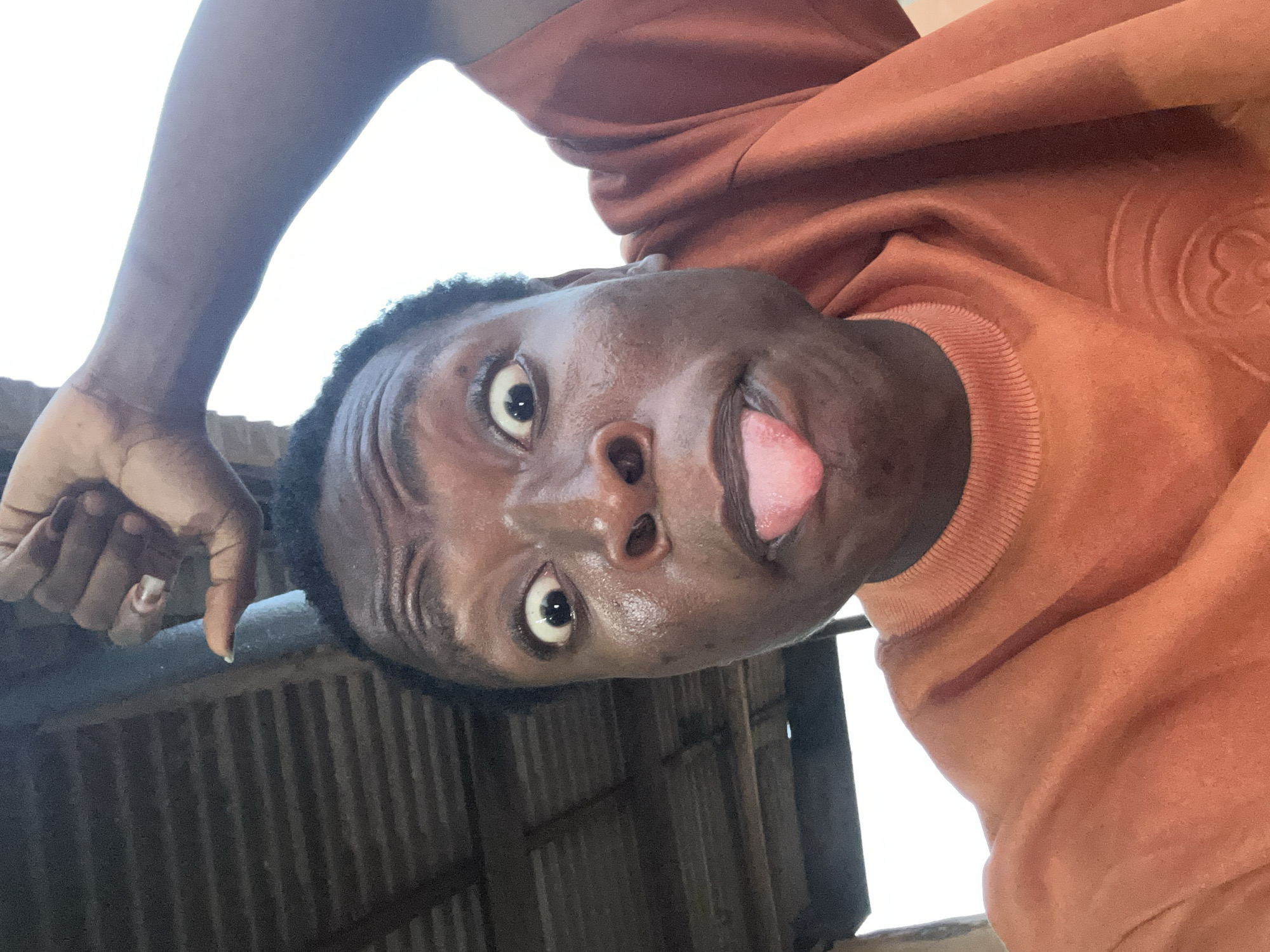 The image shows a person with dark skin wearing an orange sweatshirt, sticking their tongue out. They are positioned in a way that suggests they are looking down at the camera. The person's left arm is visible in the upper left corner of the image. The background appears to be a metal structure with a corrugated texture. The location is Unknown, Unknown.