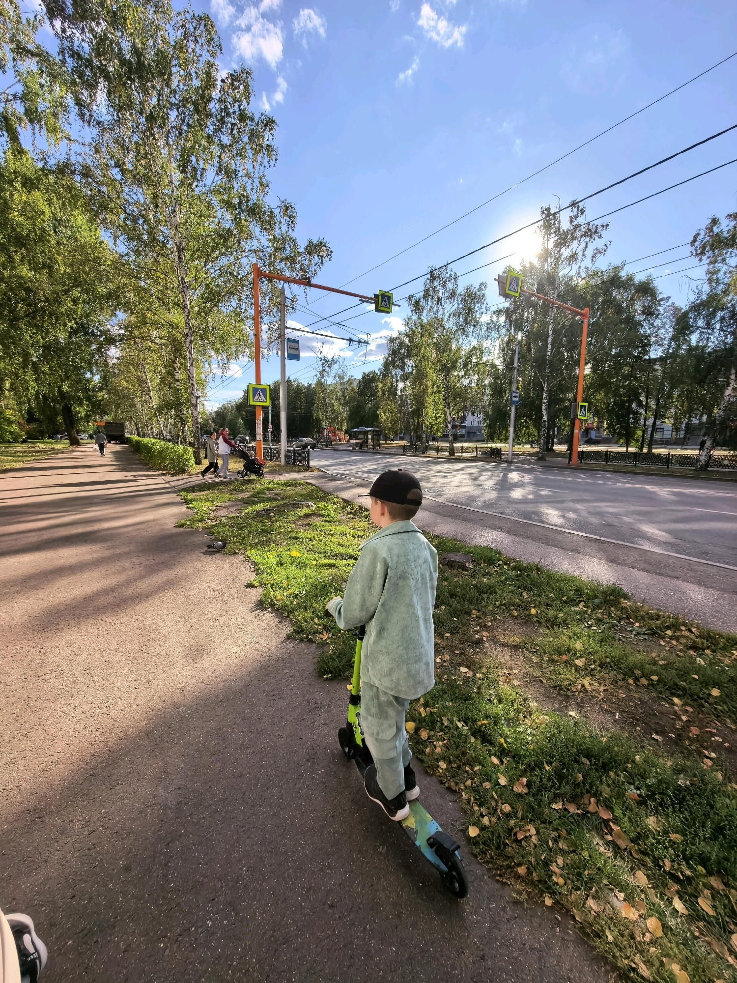 The image depicts a street scene in Sterlitamak, Russia. In the foreground, a young boy wearing a light green tracksuit and a black baseball cap rides a green and blue scooter along a paved sidewalk. He is facing away from the camera, looking towards a street with traffic and overhead wires.
The sidewalk is bordered by a strip of grass with fallen autumn leaves. Beyond the grass is a paved road with cars and what appear to be bus stops. Traffic lights and pedestrian crossing signs are visible on orange poles along the street. Trees with green and yellowing foliage line the sidewalk and the opposite side of the road. The sky is blue with scattered white clouds and bright sunlight, suggesting it is daytime. In the background, buildings are visible. A pigeon is present on the grass near the sidewalk. Further down the sidewalk, a woman pushes a stroller, accompanied by another person. Another individual is walking in the distance. The overall atmosphere suggests a typical urban environment during a sunny autumn day.