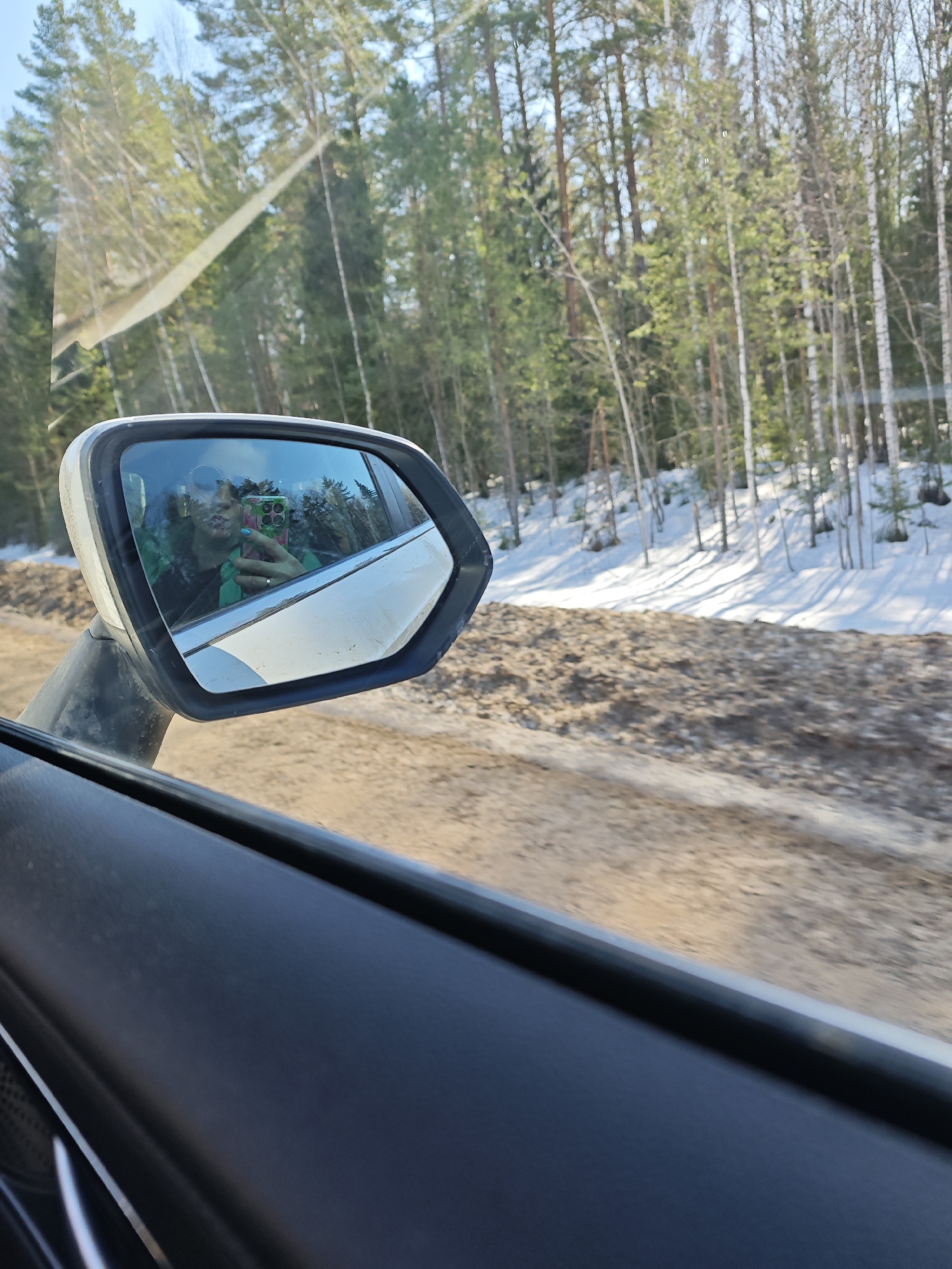 The image shows the passenger side view from inside a car. The side mirror reflects a woman taking a selfie with a cell phone; she wears a green jacket. Outside the car, a dirt road is visible alongside a snowy forest with tall pine trees and birch trees. The location is likely in a wooded area near Dubovaya, Russia, based on the presence of birch and pine trees alongside snow. The car appears to be in motion.