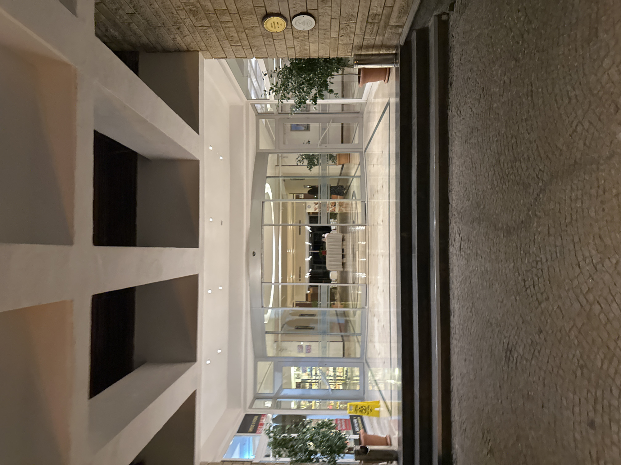 The image shows the entrance to a building, likely a hotel or resort, in Marmaris, Turkey. The entrance features large glass doors, including a revolving door in the center, and a straight sliding door to the right. There are steps leading up to the entrance, and the ground in front of the steps is paved with cobblestones.
Inside the building, a long table draped with a white tablecloth is visible, suggesting a dining or reception area. Two potted plants are placed outside the entrance, one on each side. The building's exterior is a combination of brickwork on the right side and a modern facade with geometric ceiling designs. Two circular plaques are mounted on the brick wall on the right. A yellow "Wet Floor" sign is placed on the left side near the entrance. The scene appears to be captured at dusk or night, with artificial lighting illuminating the area.
