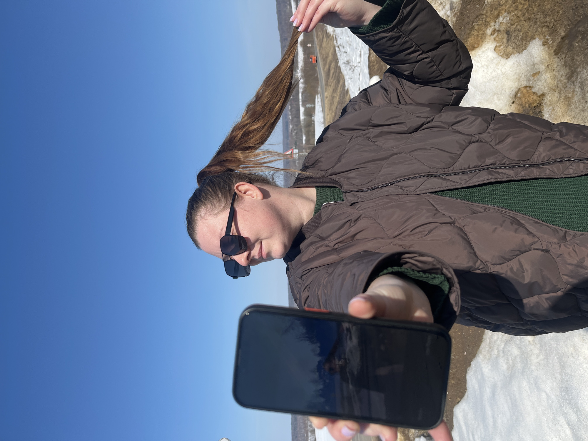 An individual stands outdoors under a clear blue sky, holding a dark-colored smartphone forward in their left hand, its screen reflecting the surrounding landscape. The individual is a female wearing dark rectangular sunglasses, a dark brown quilted jacket over a dark green knitted sweater, and has brown hair tied in a high ponytail. Her right hand is seen touching a section of her ponytail. She is looking towards her right. The background, identified as Zubovo, Russia, features a wide landscape with patches of melting snow on the ground in the foreground and on distant rolling hills. A paved road or highway, on which an orange-red vehicle is visible in the distance, winds through the valley. A portion of a white road sign with a red border is partially visible behind the individual's head.