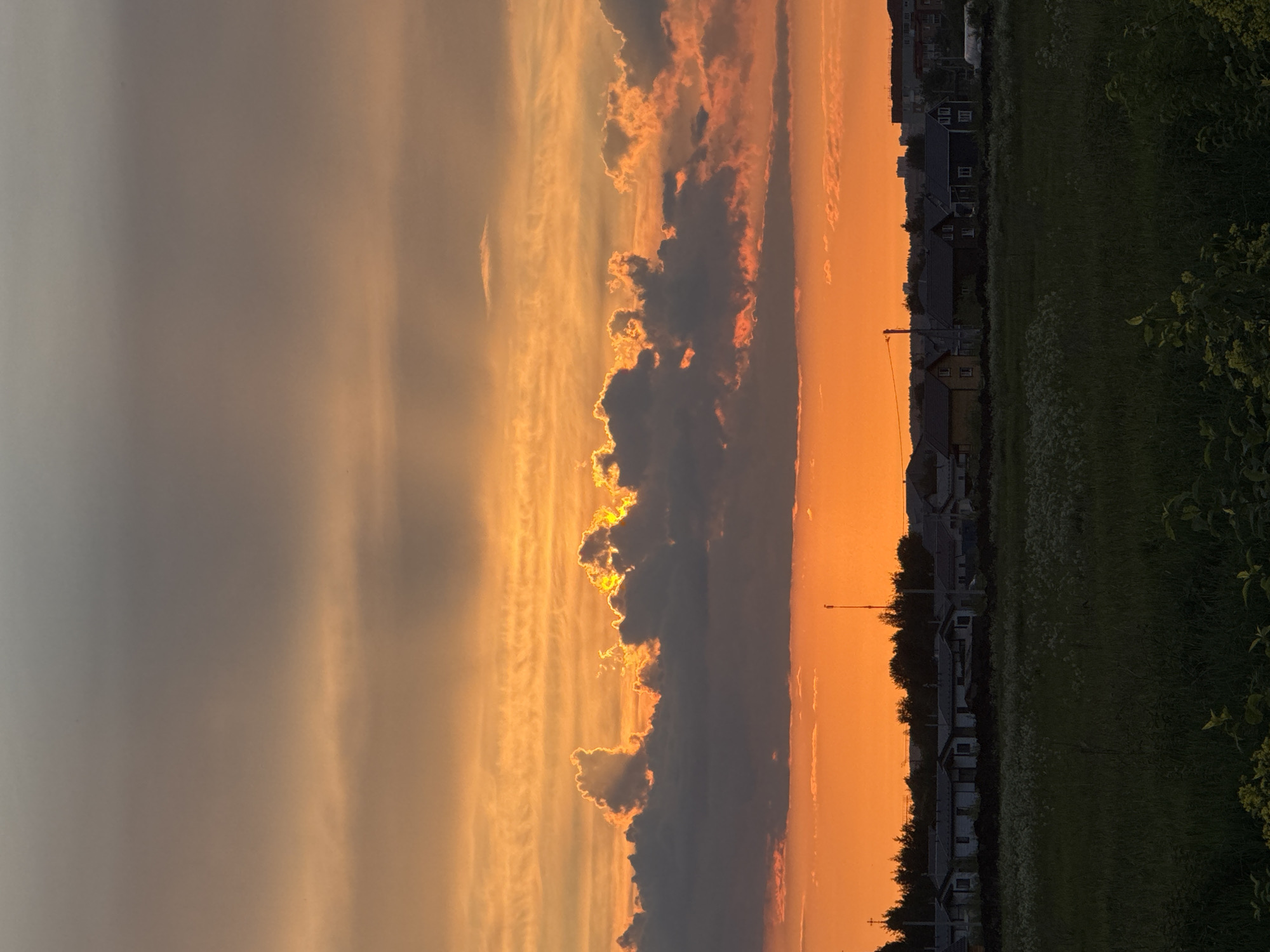 The image captures a dramatic sunset over a rural or suburban landscape in Горный, Russia. The scene is dominated by a vibrant, multi-hued sky and silhouetted clouds.
A prominent band of dark, irregularly shaped clouds stretches across the middle of the sky. These clouds are intensely backlit by the setting sun, causing their edges to glow with bright yellow and fiery orange light, creating a striking contrast against their darker bodies. Above this central cloud formation, the sky transitions from a textured warm orange to a softer, muted grey towards the upper left. Below the main cloud band, the sky is a deep, intense orange, indicating the sun's position near or just below the horizon.
Along the horizon line, a dark silhouette of residential houses and trees is visible, stretching from left to right. These buildings appear to be single or two-story homes, standing out sharply against the brilliant orange backdrop. A tall, slender structure, possibly a utility pole or antenna, can also be seen among the houses. The foreground consists of a dark field, likely covered in grass and other vegetation, with some lighter green details discernible on the right side.
The time of day is clearly sunset, characterized by the warm, glowing colors. No specific activity or visible text is present in the image. The overall impression is one of natural beauty and serenity, highlighting the dramatic interplay of light and shadow during dusk.