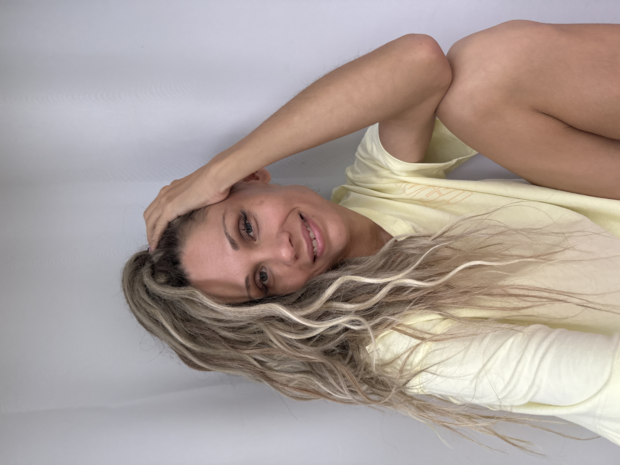 A light-skinned woman with blonde dreadlocks is sitting against a white background in Zhukovo, Russia. She is wearing a light yellow t-shirt and has her right hand on top of her head. Her left arm is bent and resting on her leg, and her left leg is bent and tucked towards her body. She is smiling and looking towards the camera. The background is a plain white wall.