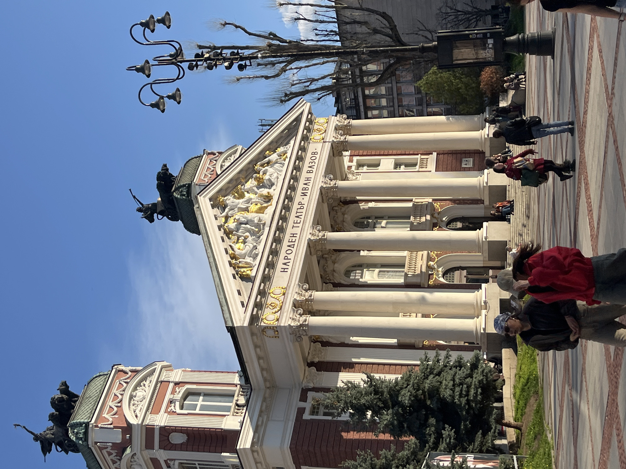 The image shows the Ivan Vazov National Theatre in Sofia, Bulgaria. The building has a classical facade with white columns, sculptures, and inscriptions. A street lamp stands to the left of the building. People are walking on the pavement in front of the theatre.