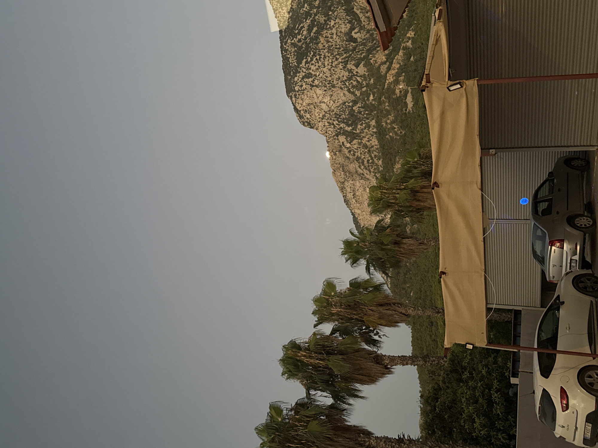 The image depicts a view of the coastline and mountains in Kemer, Turkey. In the foreground, a beige awning covers a structure with rolled-down gray shutters, suggesting it might be a shop or an establishment. Two cars are parked in front of the shutters: a white Renault Clio on the left and a silver sedan on the right. Behind the awning, a row of palm trees stands against the backdrop of a rugged, tree-covered mountain range. The moon is visible on the face of the mountain, just peeking over the crest. The sky is a pale gray, indicating either early morning or late evening light. The overall scene conveys a tranquil, coastal atmosphere.